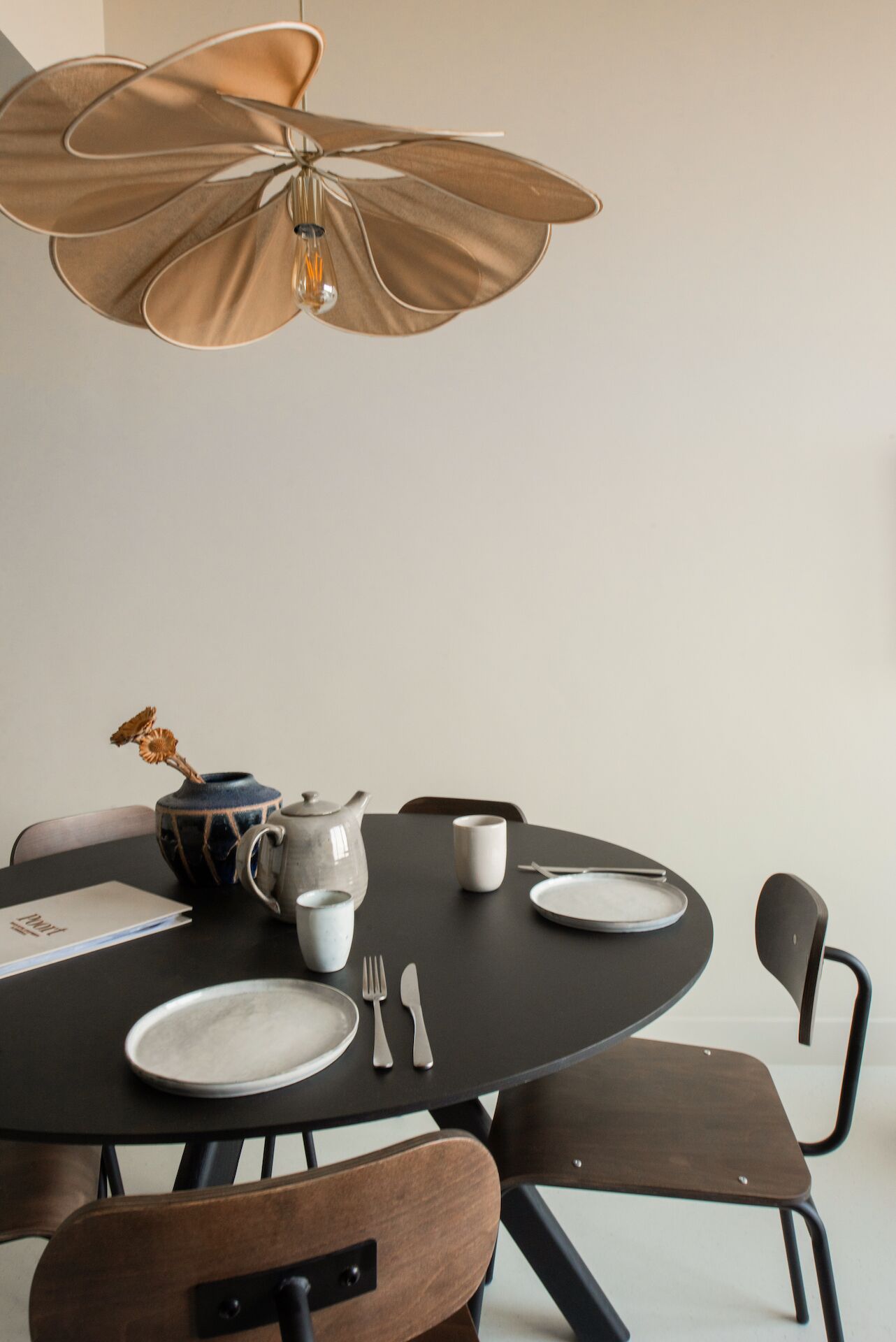 Une table de salle à manger ronde, recouverte d’une nappe noire, avec un vase contenant des fleurs séchées, une théière, une tasse et des couverts. Un luminaire suspendu se trouve au-dessus de la table.