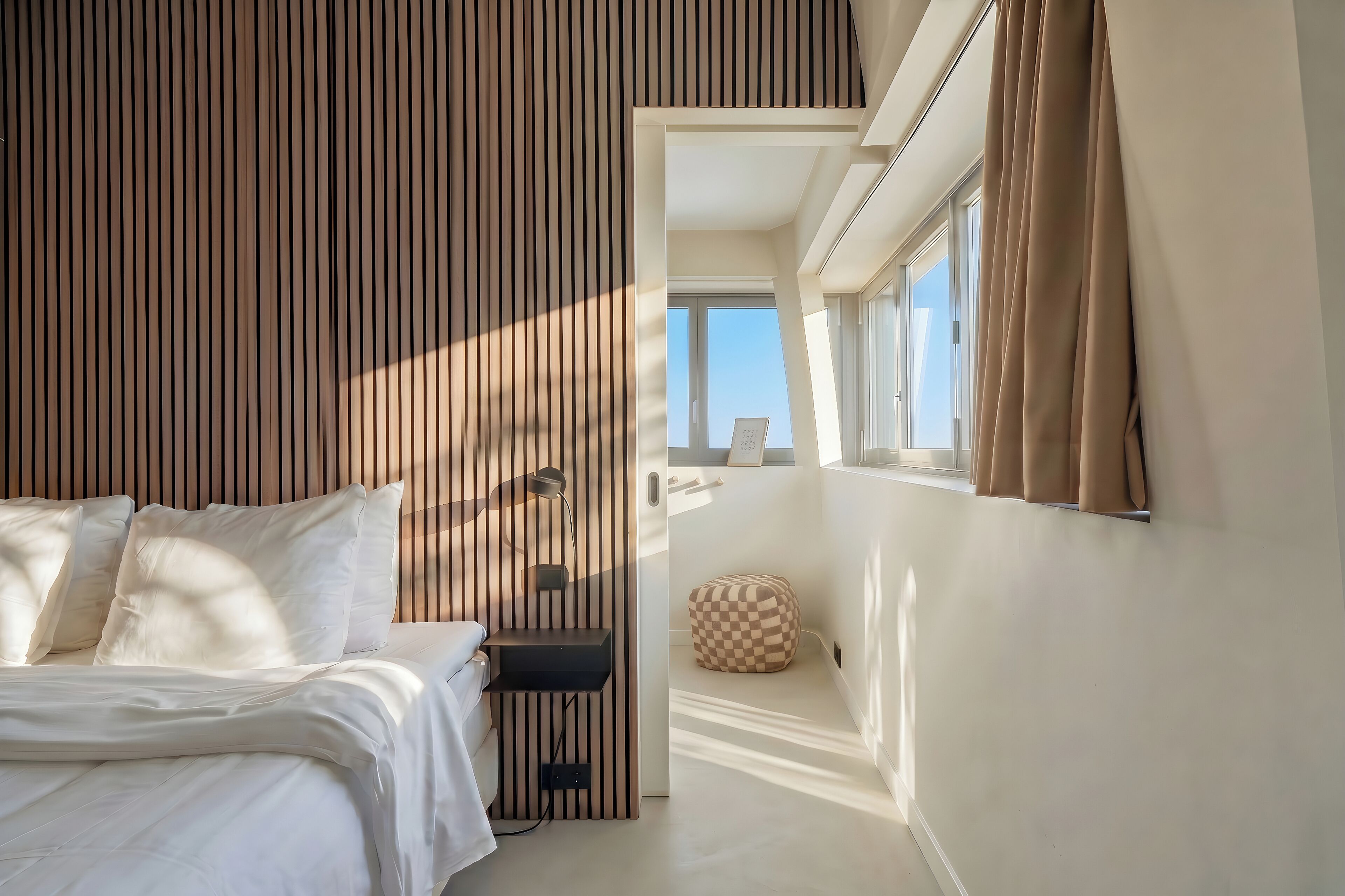Un dormitorio con cama, mesita de noche, lámpara, ventana con cortinas y vista al cielo.