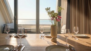 Een eettafel met lege borden en wijnglazen, een vaas met bloemen en uitzicht op zee door grote ramen.