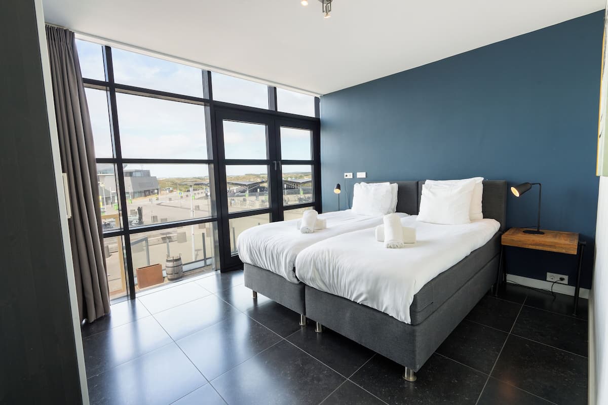Habitación de hotel moderna con una cama grande, una mesita de noche y vistas de la ciudad a través de amplias ventanas.