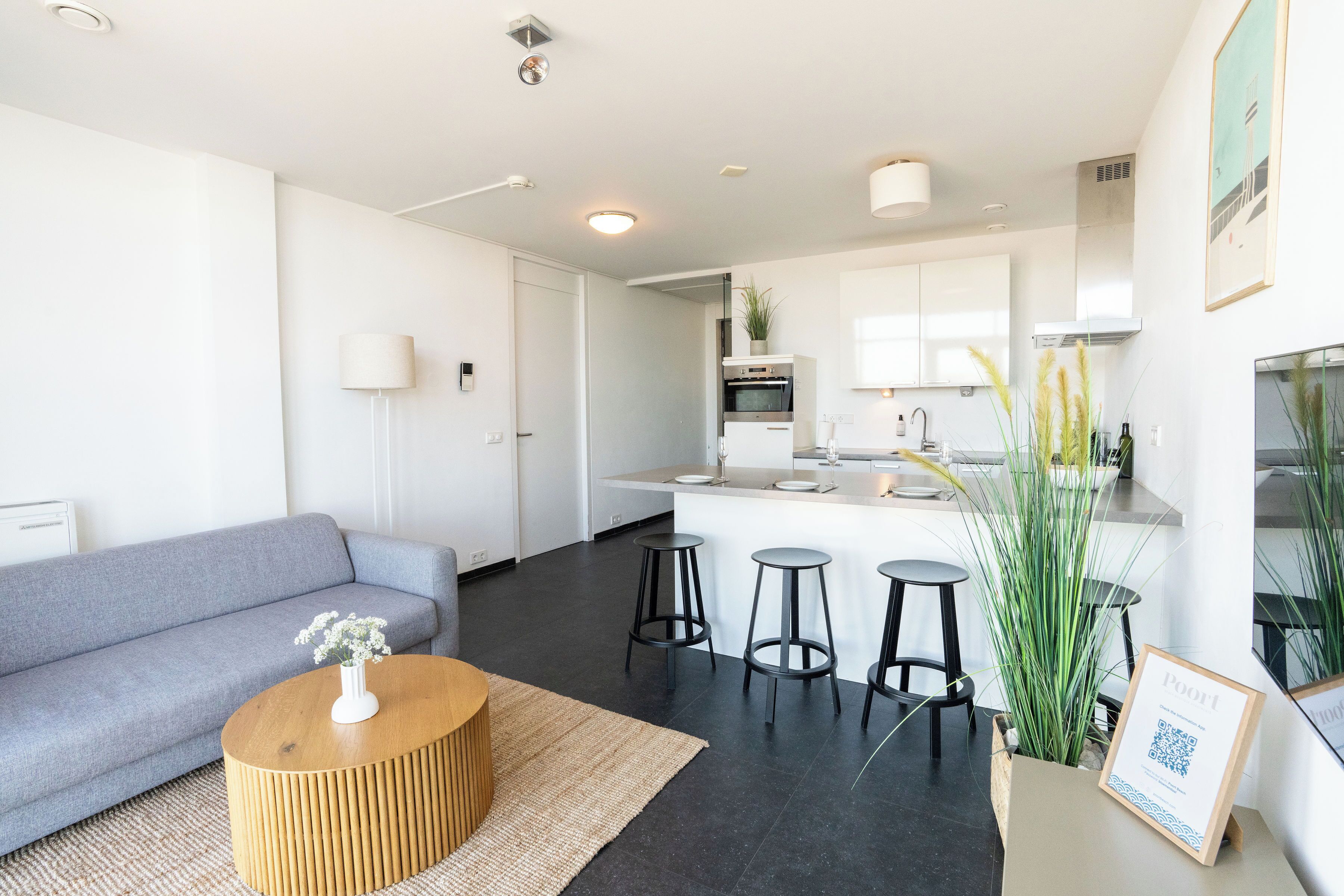 Un apartamento moderno con un sofá gris, una mesa de centro redonda de madera, y una zona de cocina con una mesa de comedor y taburetes.