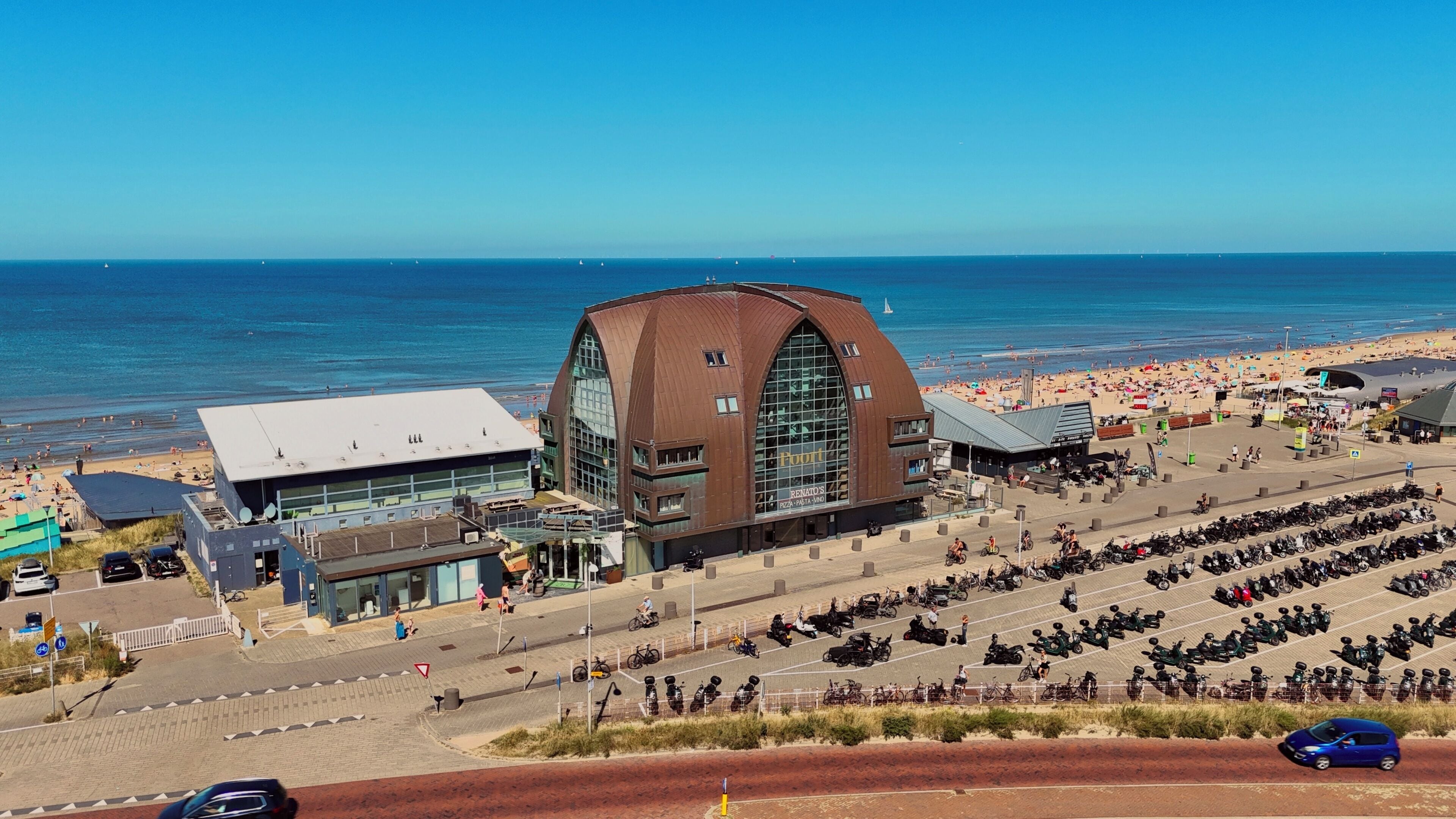 A coastal building with a large arched entrance, a paved area with parked scooters, and a beach in the background.