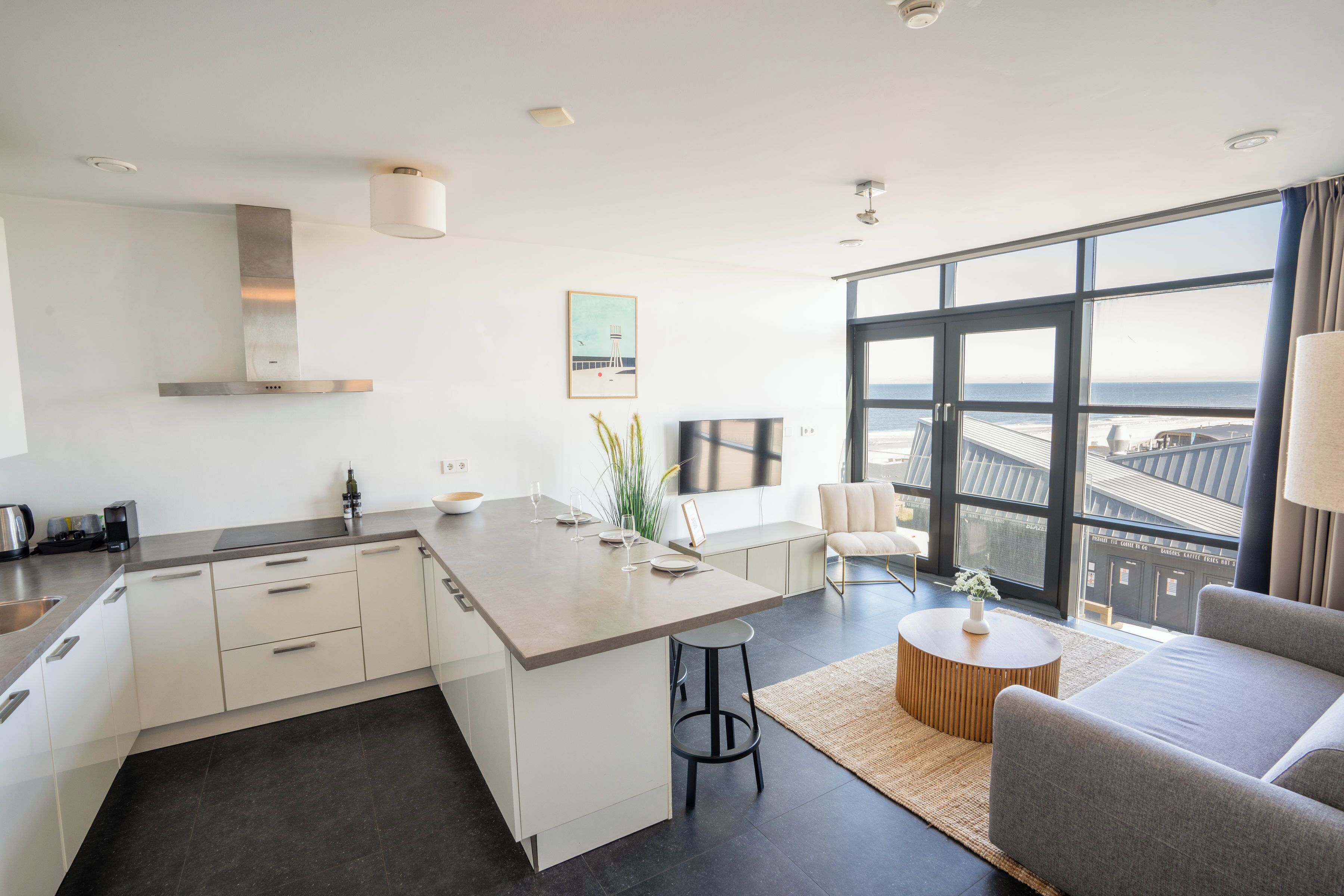 A modern kitchen with a central island, a dining area with a sofa and a round coffee table, and a large window offering a view of the cityscape.