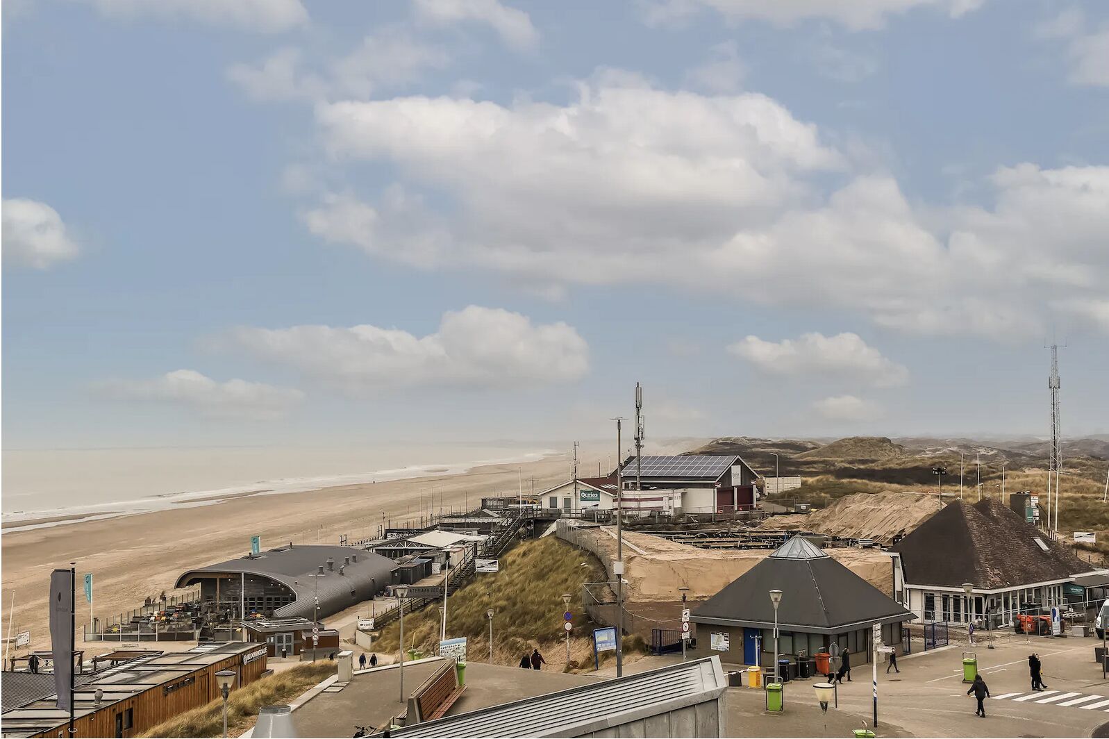 A coastal area with a beach, a small building with a sign, and a large structure with a sloped roof.