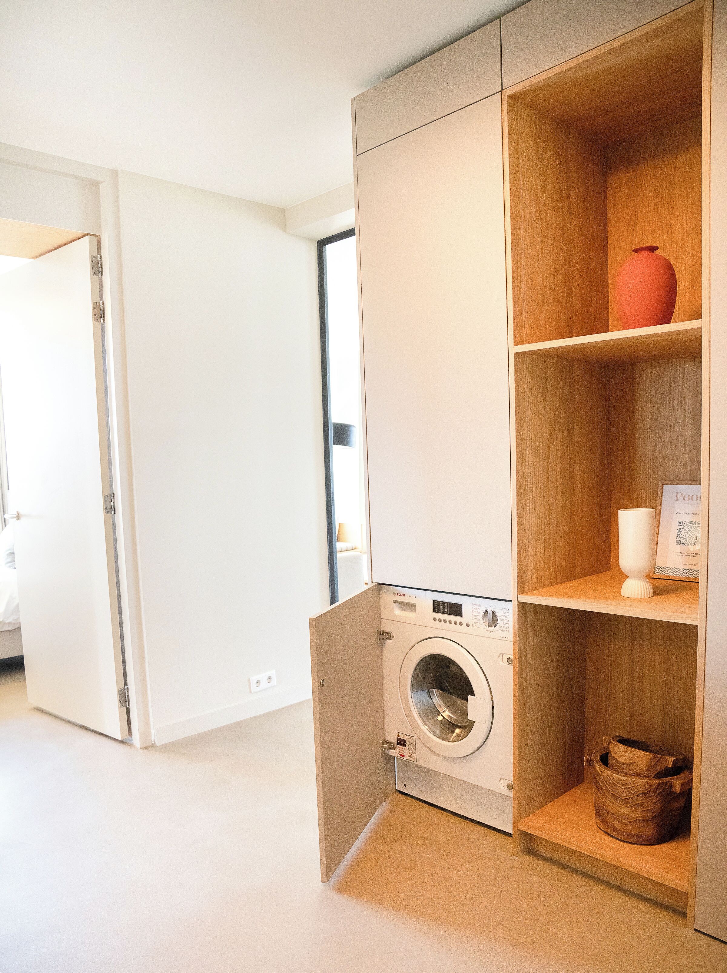 Une petite buanderie équipée d’un lave-linge Bosch, d’étagères en bois et d’une porte donnant sur une autre pièce.