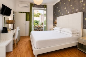 A hotel room with a bed, a desk, a chair, a TV, and a balcony with a view of greenery. - B&B Pellicano Guest House (Reggio Calabria)