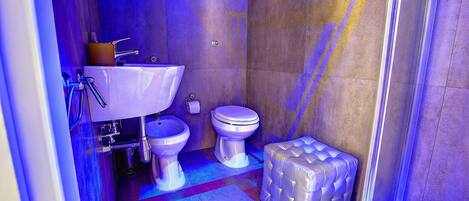 A bathroom with a white sink, toilet, and bidet, a small cushioned bench, and a shower area.