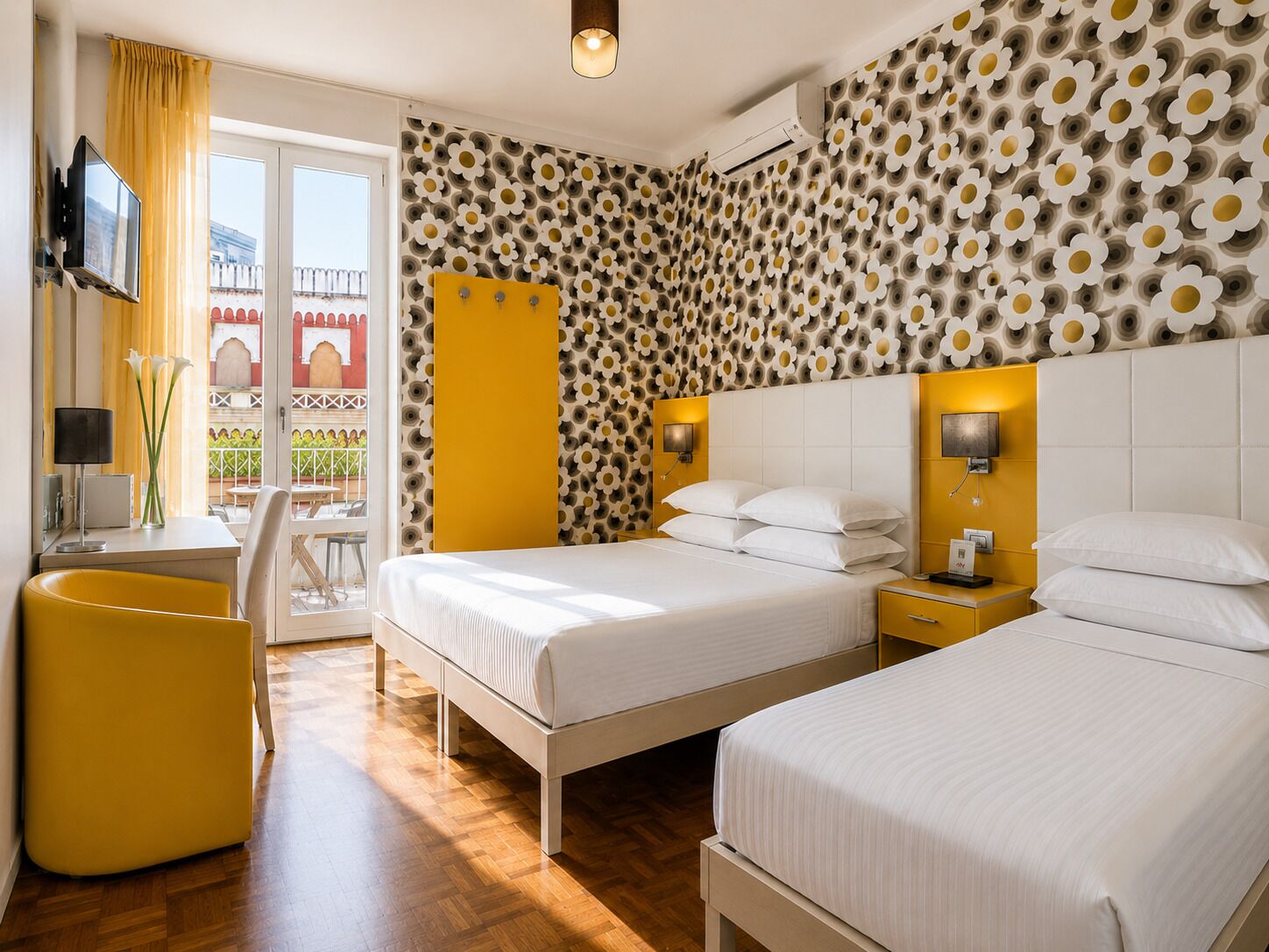 Quarto de hotel com duas camas, uma mesa de trabalho com cadeira, um banquinho amarelo e um papel de parede estampado.