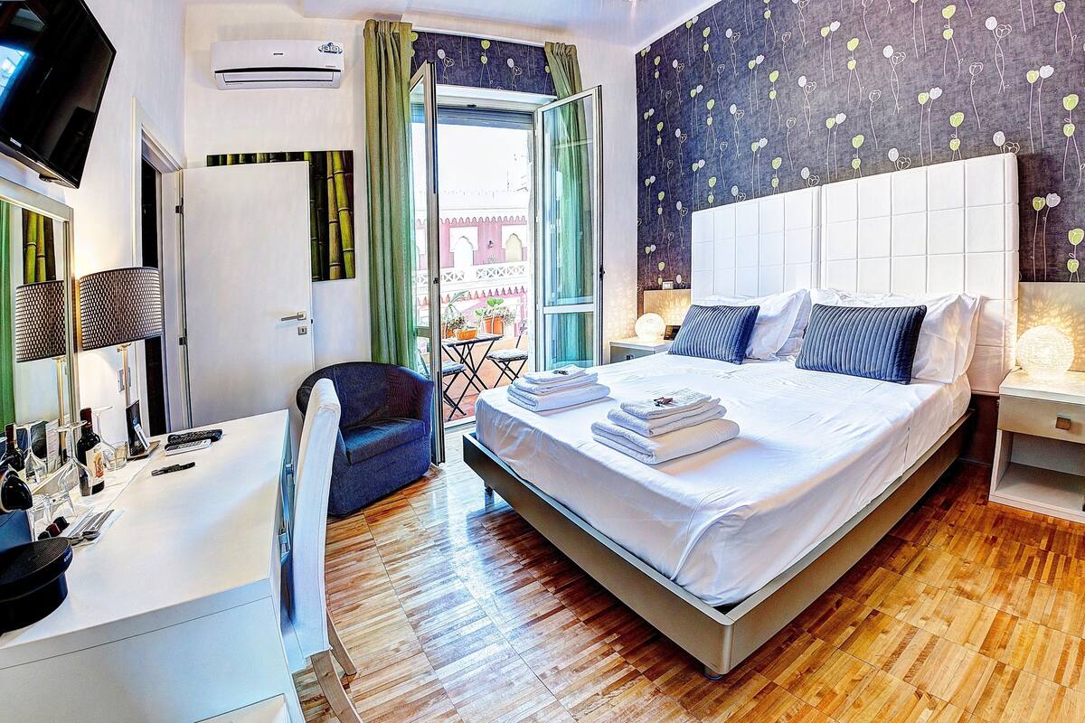 A modern hotel room with a large bed, a desk with a computer, a chair, and a view of the outside balcony.
