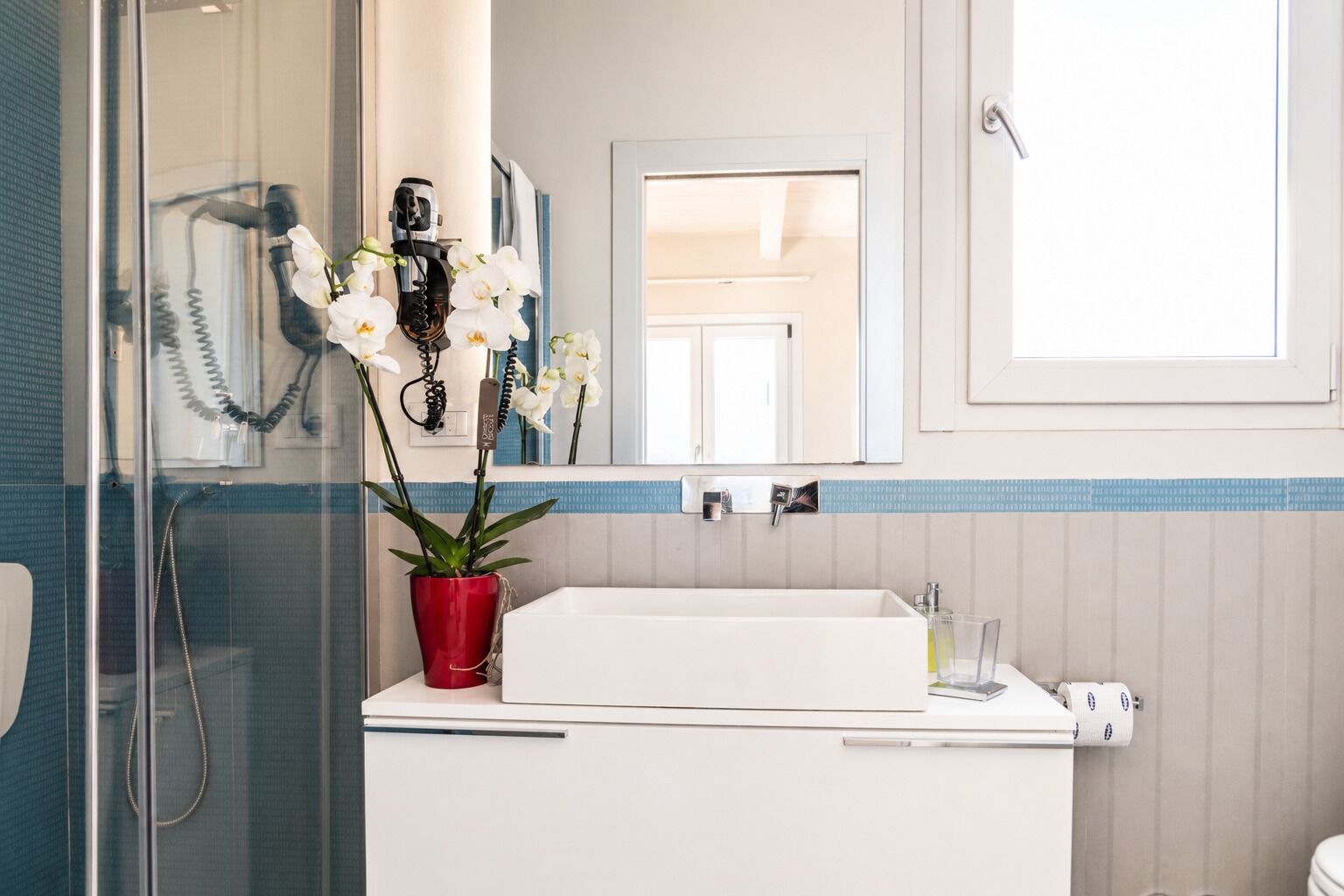 A bathroom with a white sink, a red vase with white flowers, a mirror, and a shower area.