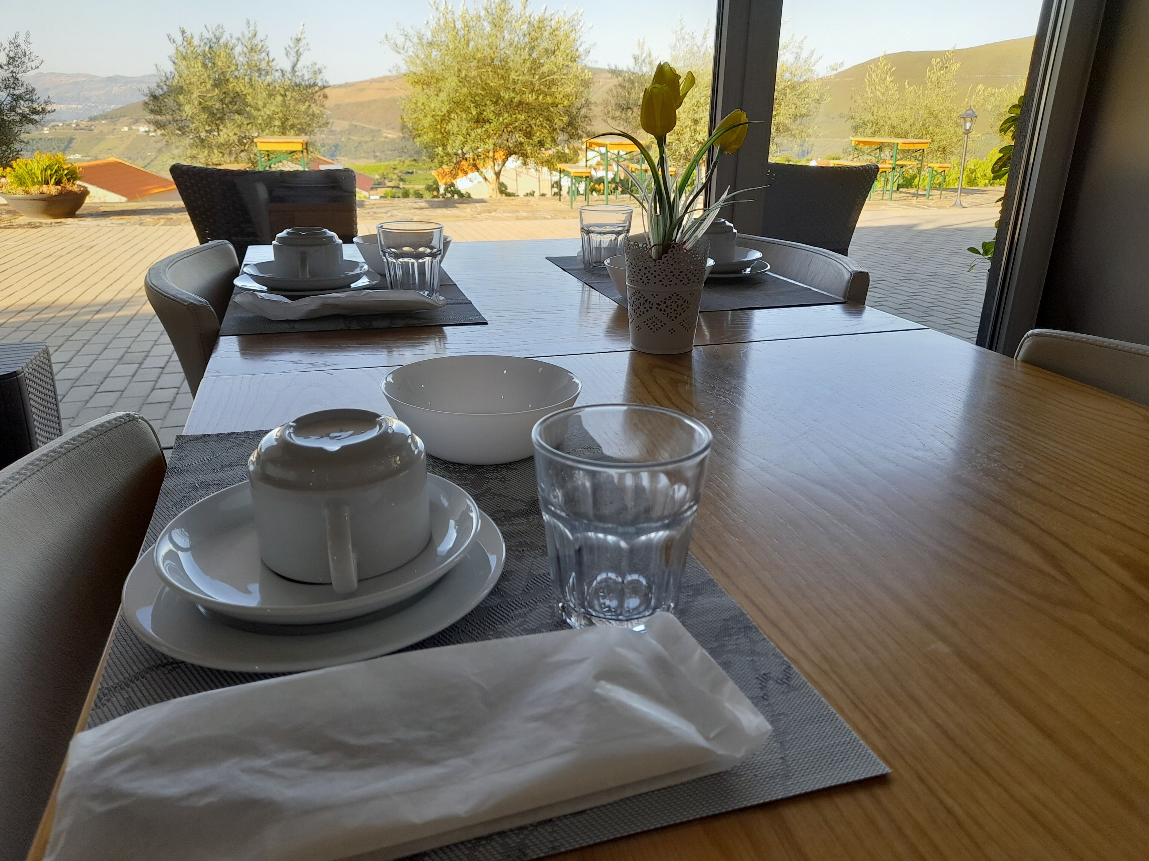 Mesa de comedor lista con vajilla blanca, vasos y servilleta, con vistas a un espacio exterior pintoresco con sillas y mesas.