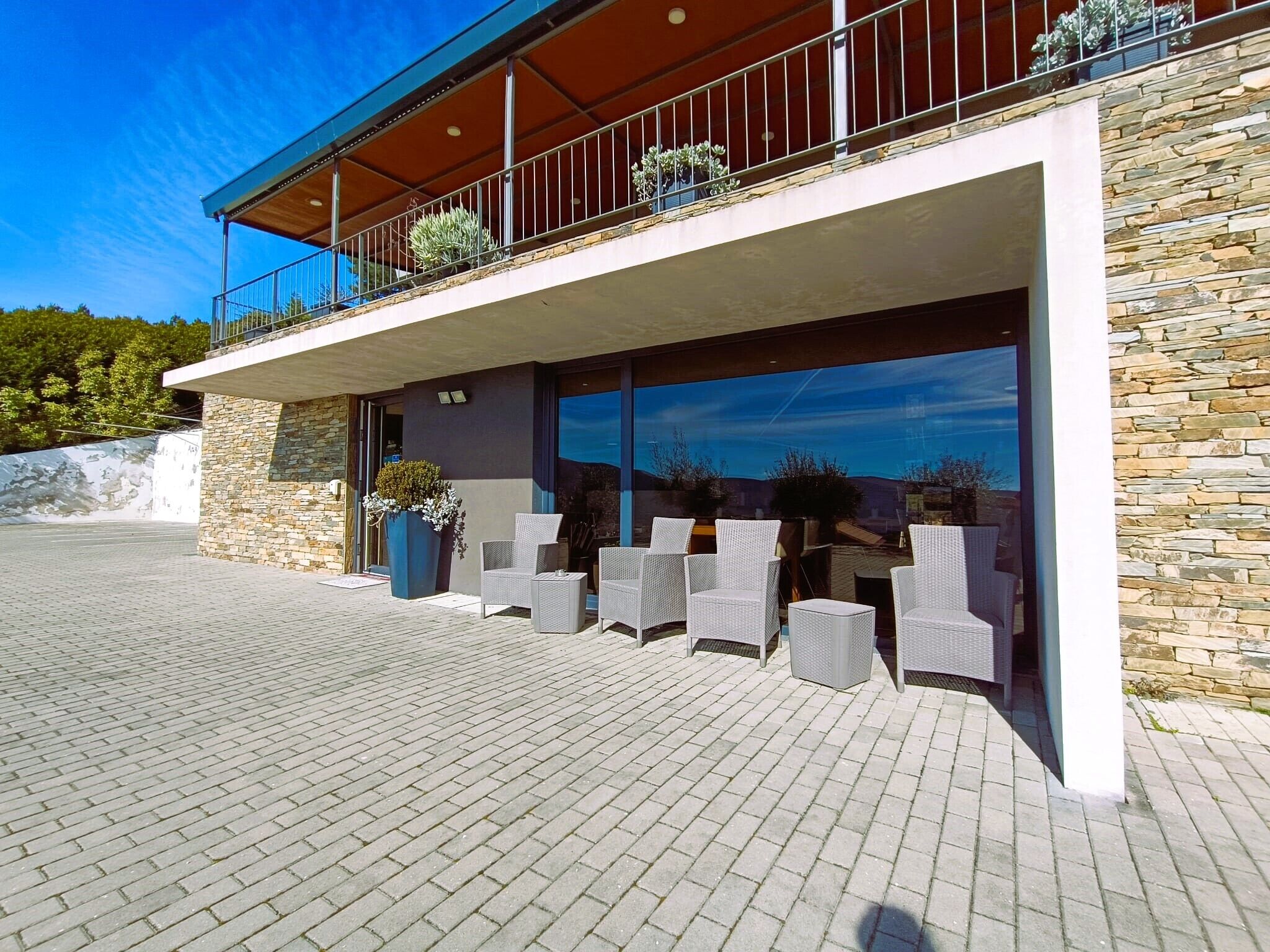 A modern building with a stone facade, a balcony with outdoor seating, and a large window reflecting the sky.