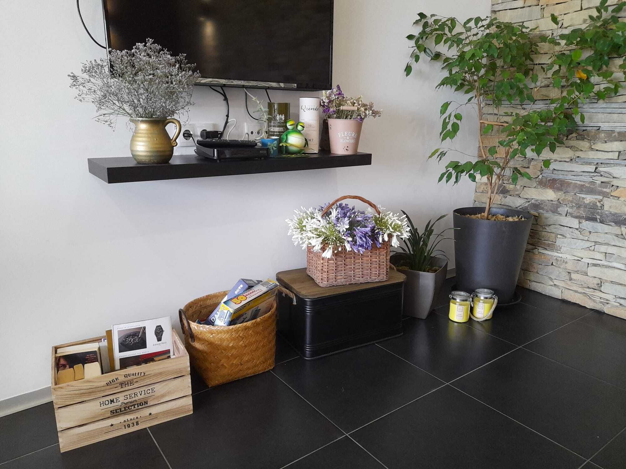 A modern living room with a flat-screen TV mounted on the wall, a stone wall, a potted plant, a shelf with decorative items, a basket of flowers, and a wooden crate with books.