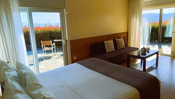 Quarto de hotel com uma cama grande, um sofá, uma mesa pequena e uma porta de vidro deslizante que dá para uma área externa com vista para montanhas e flores coloridas.