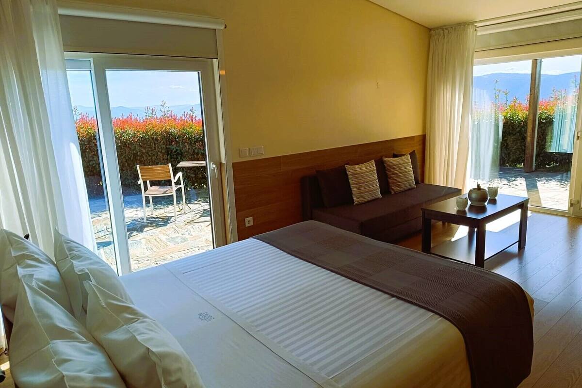 A hotel room with a large bed, a sofa, a small table, and a sliding glass door leading to an outdoor area with a view of mountains and colorful flowers.