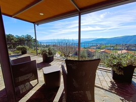 Un espacio de estar al aire libre techado con muebles de mimbre y plantas en macetas, con vistas a un paisaje pintoresco con casas y montañas.