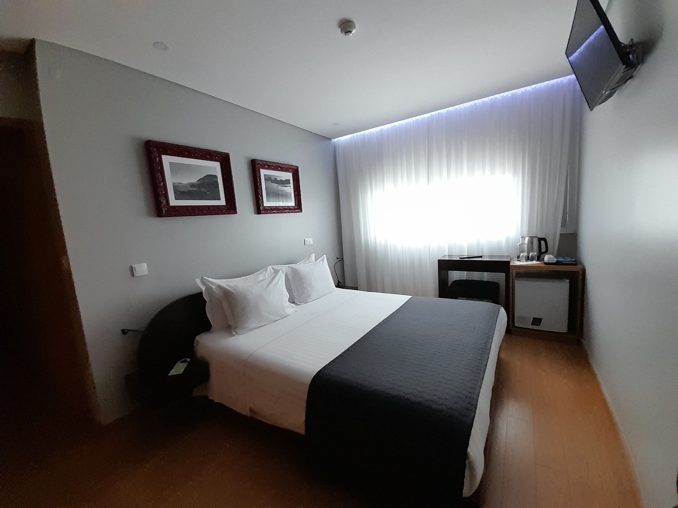 A hotel room with a bed, a desk with a chair, a TV mounted on the wall, and two framed pictures on the wall.