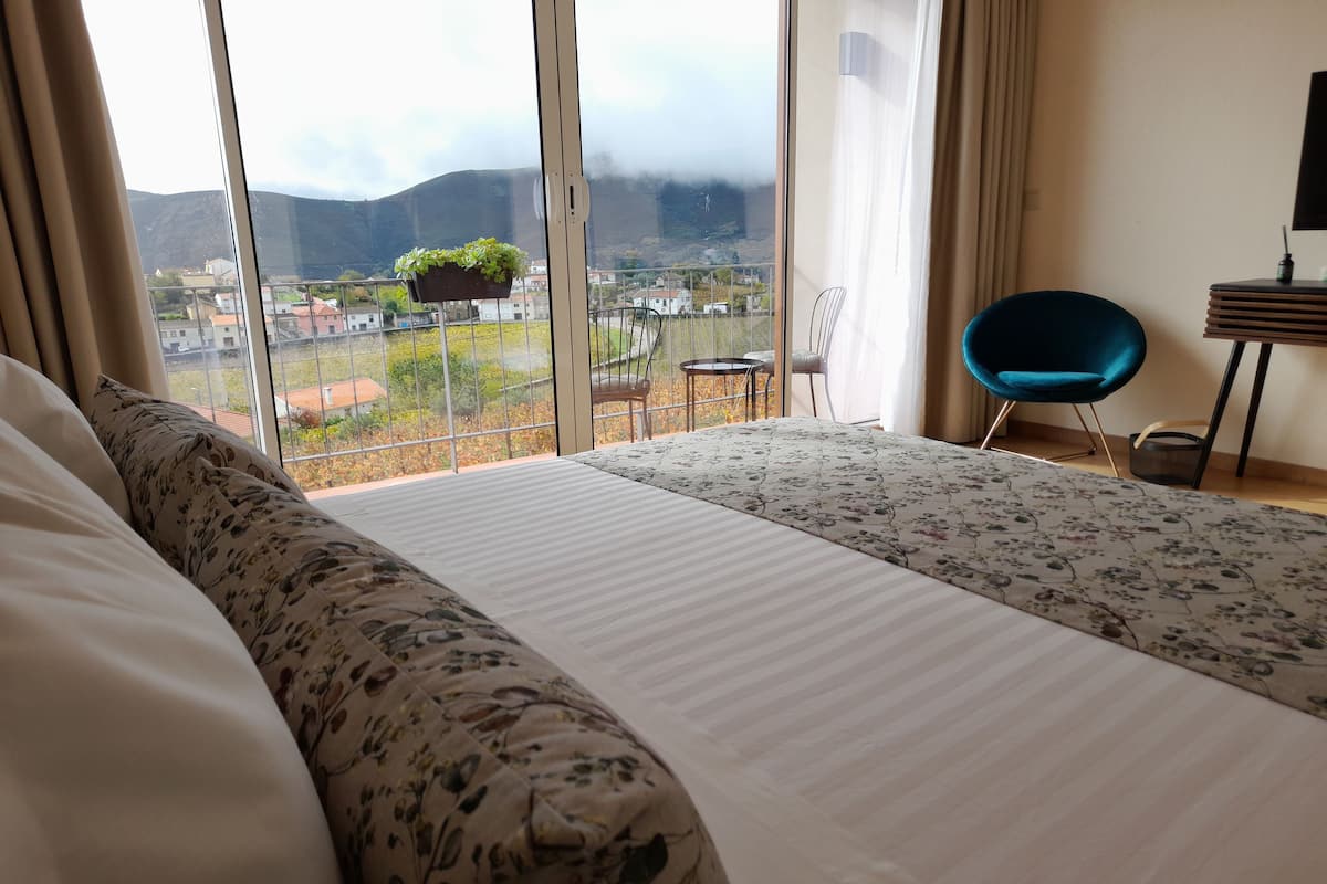 A hotel room with a large bed, a view of the city and mountains, a small balcony with a chair, and a desk with a lamp.