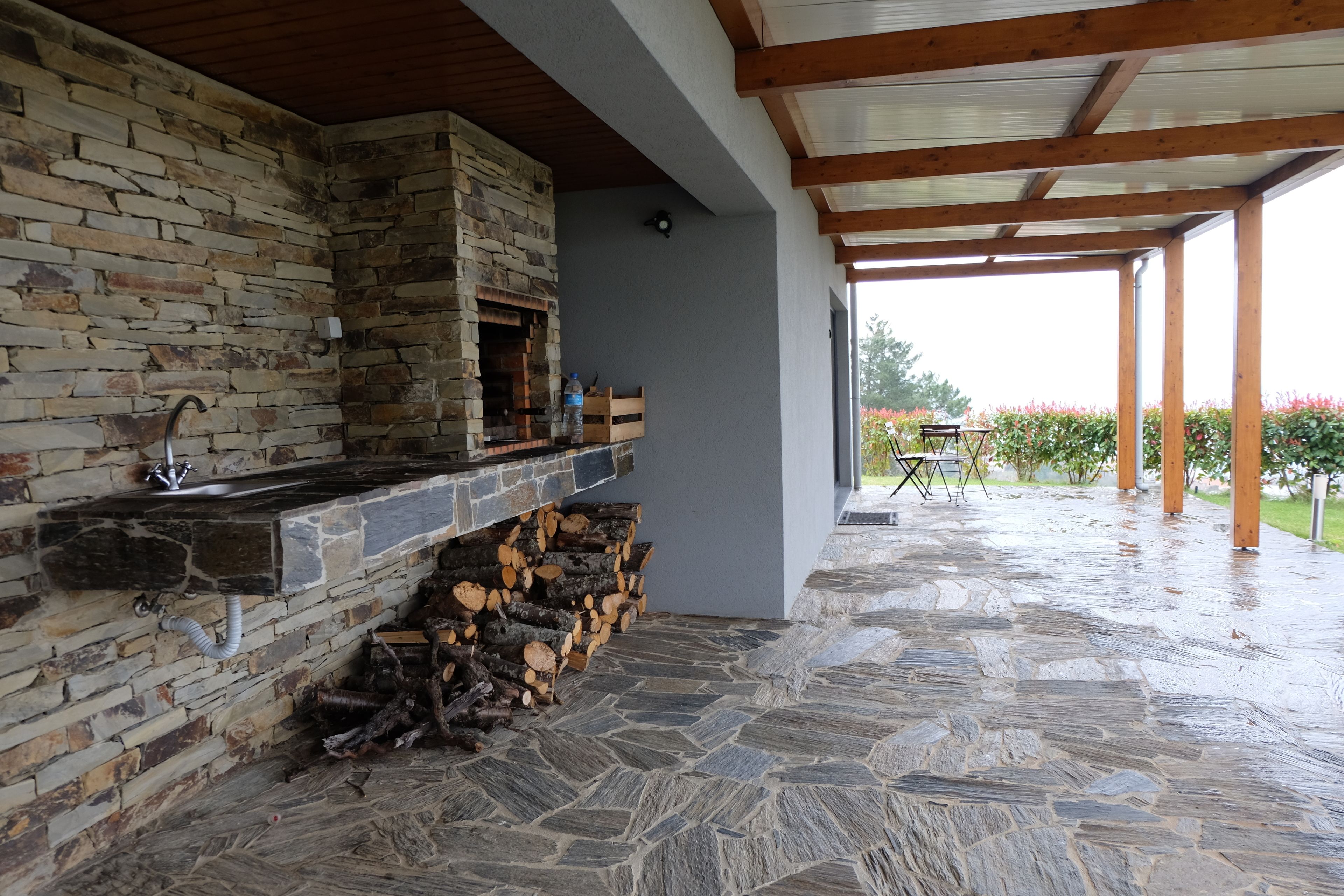 Una chimenea de piedra con un lavamanos, leña apilada y un patio cubierto con vistas a un jardín.