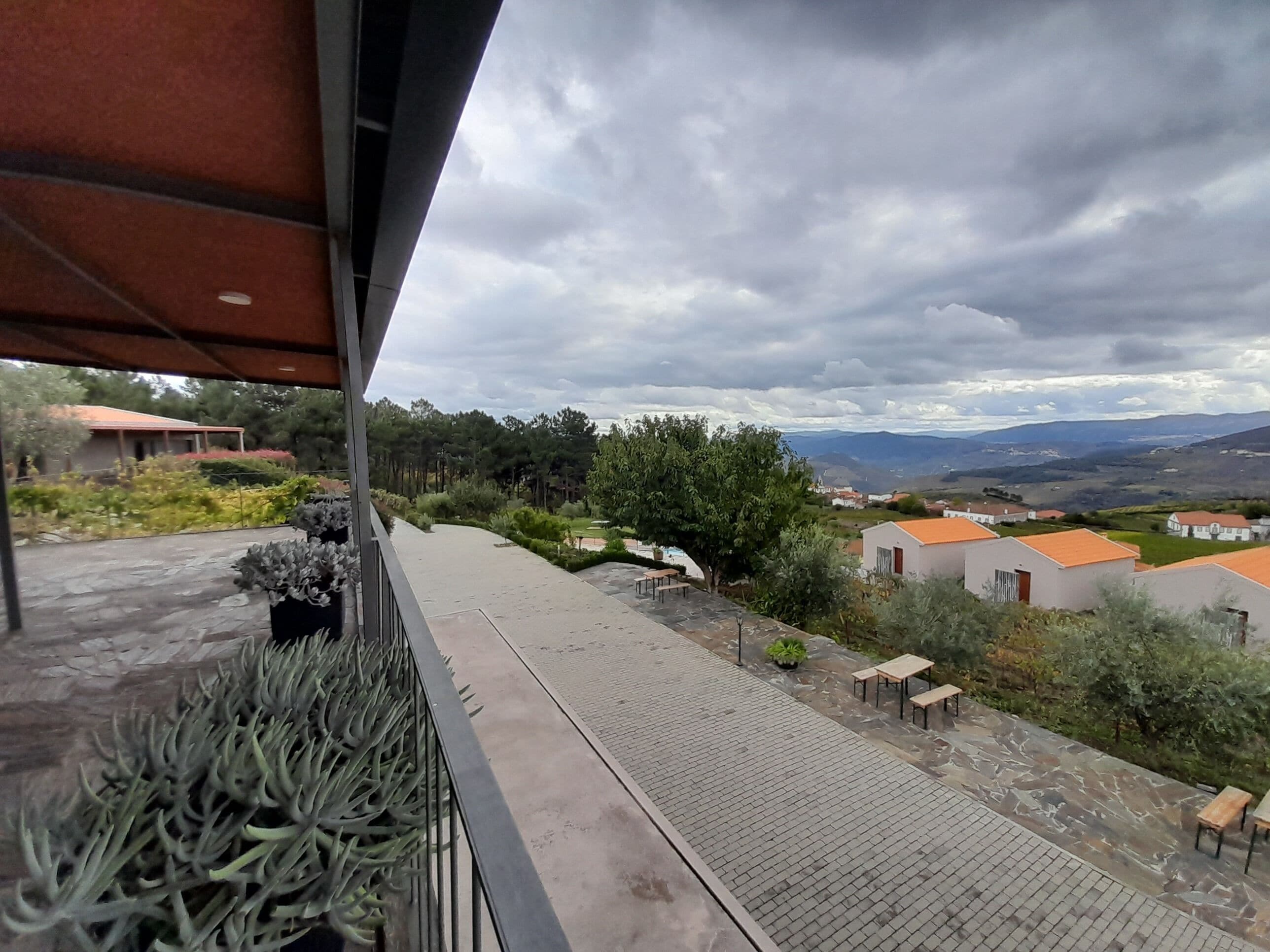 A balcony with a view of a residential area, a paved walkway, and a row of houses with orange roofs.
