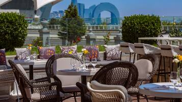 An outdoor seating area with wicker chairs and tables, a city skyline in the background, and a clear sky.