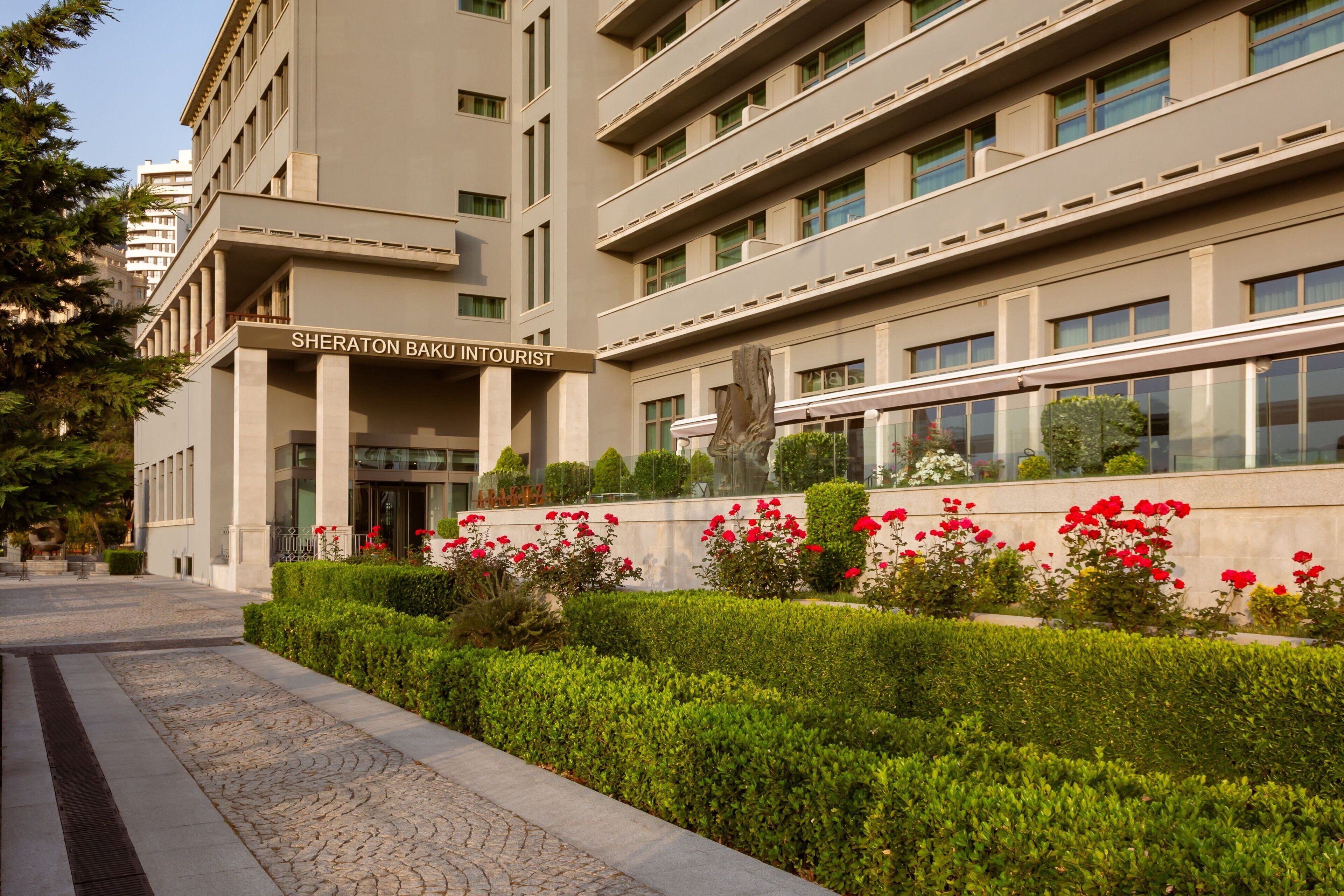 Un bâtiment Sheraton doté d’une façade moderne, d’une entrée imposante et d’un aménagement paysager bien entretenu, agrémenté de fleurs rouges.