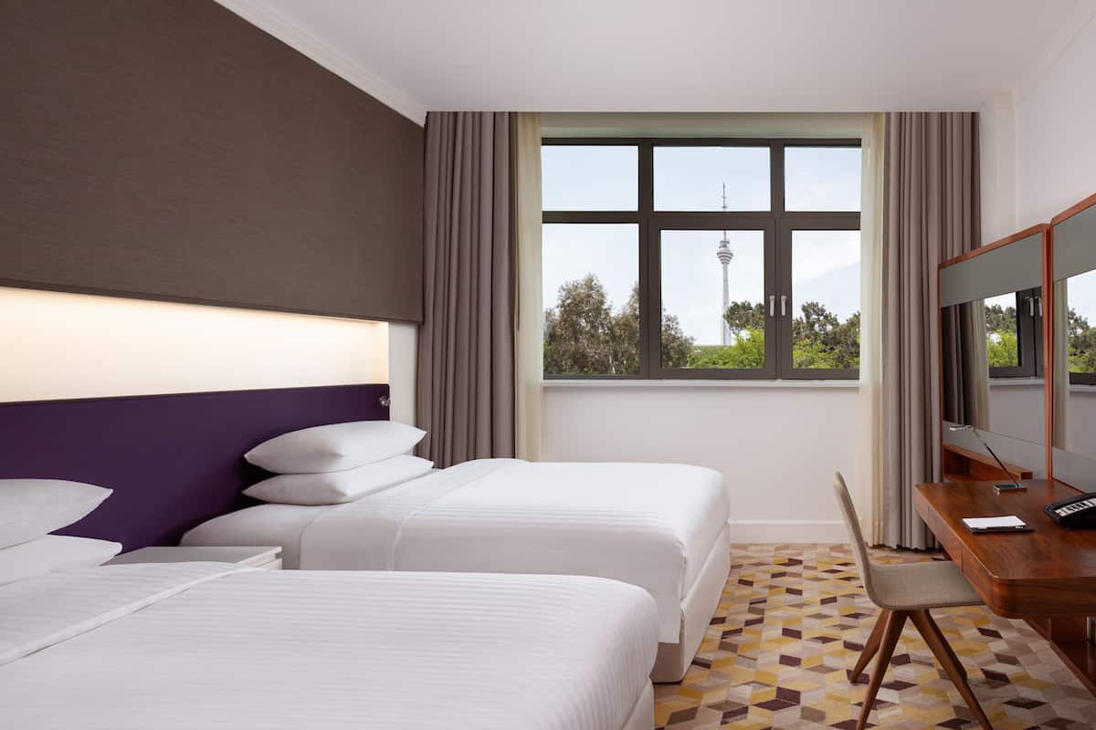 A hotel room with two beds, a desk, a chair, and a large window with a view of a tower.