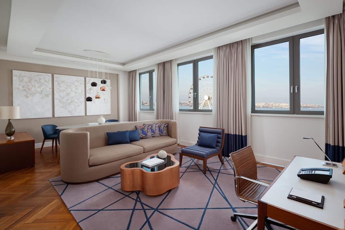 A modern hotel room with a large window, a sofa, a coffee table, a desk, and a chair.