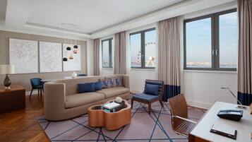 A modern hotel room with a large window, a sofa, a coffee table, a desk, and a chair.