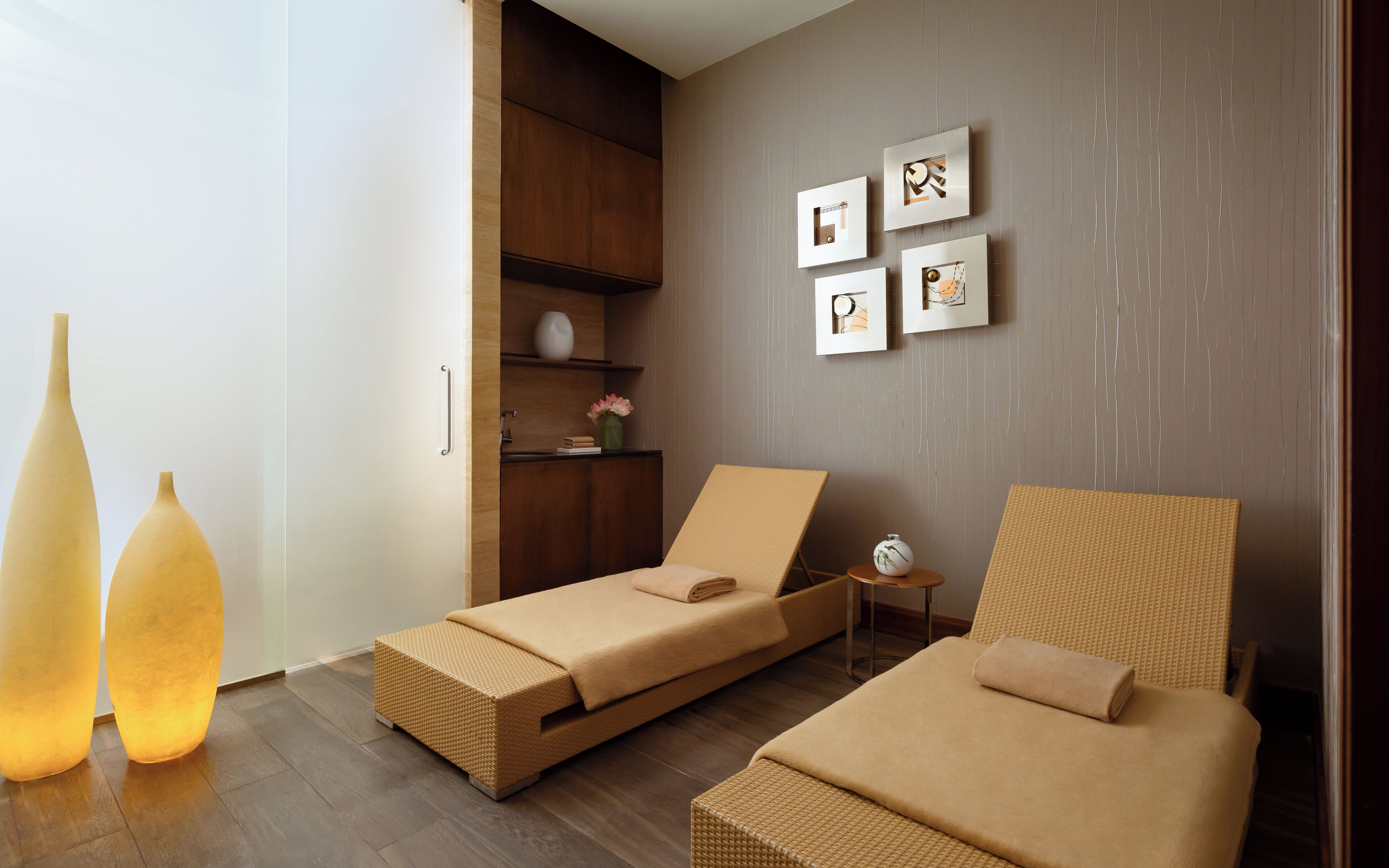 A modern hotel room with two single beds, a wooden wardrobe, a small side table, and wall-mounted artwork.