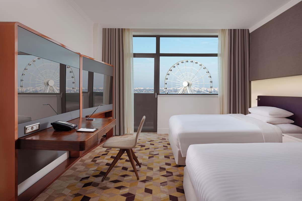A hotel room with two beds, a desk with a computer, and a large window with a view of a Ferris wheel.