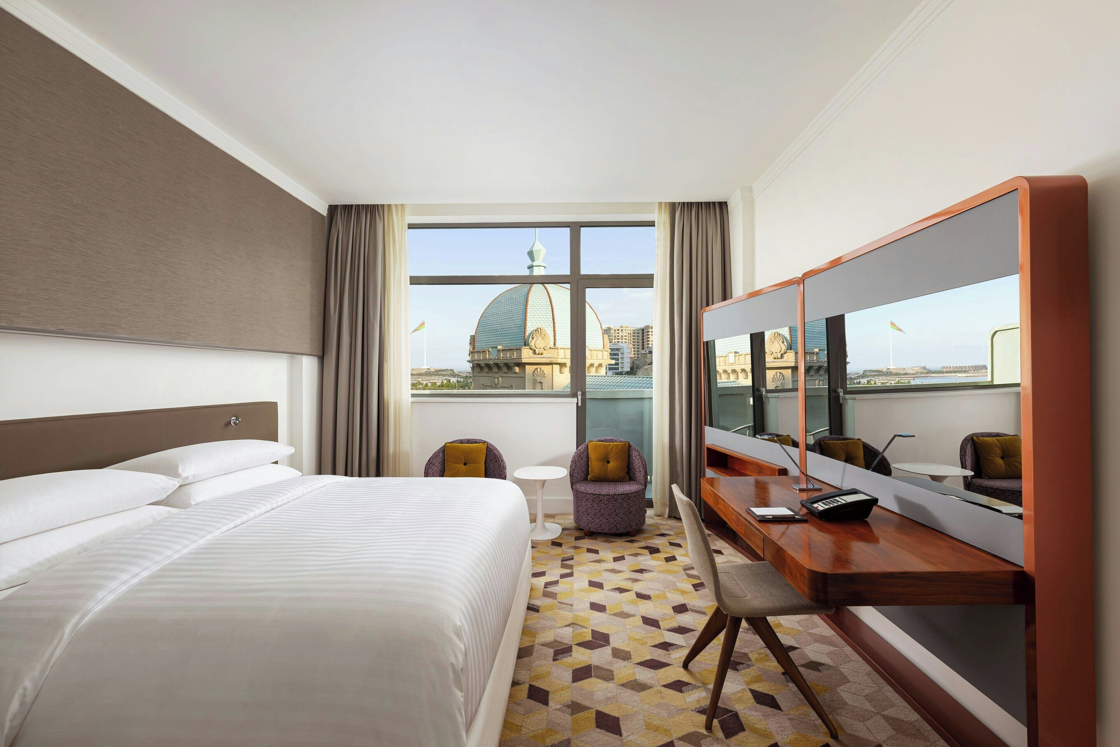 A hotel room with a large bed, a desk with a chair, a small table, and a large window offering a view of a dome and buildings.