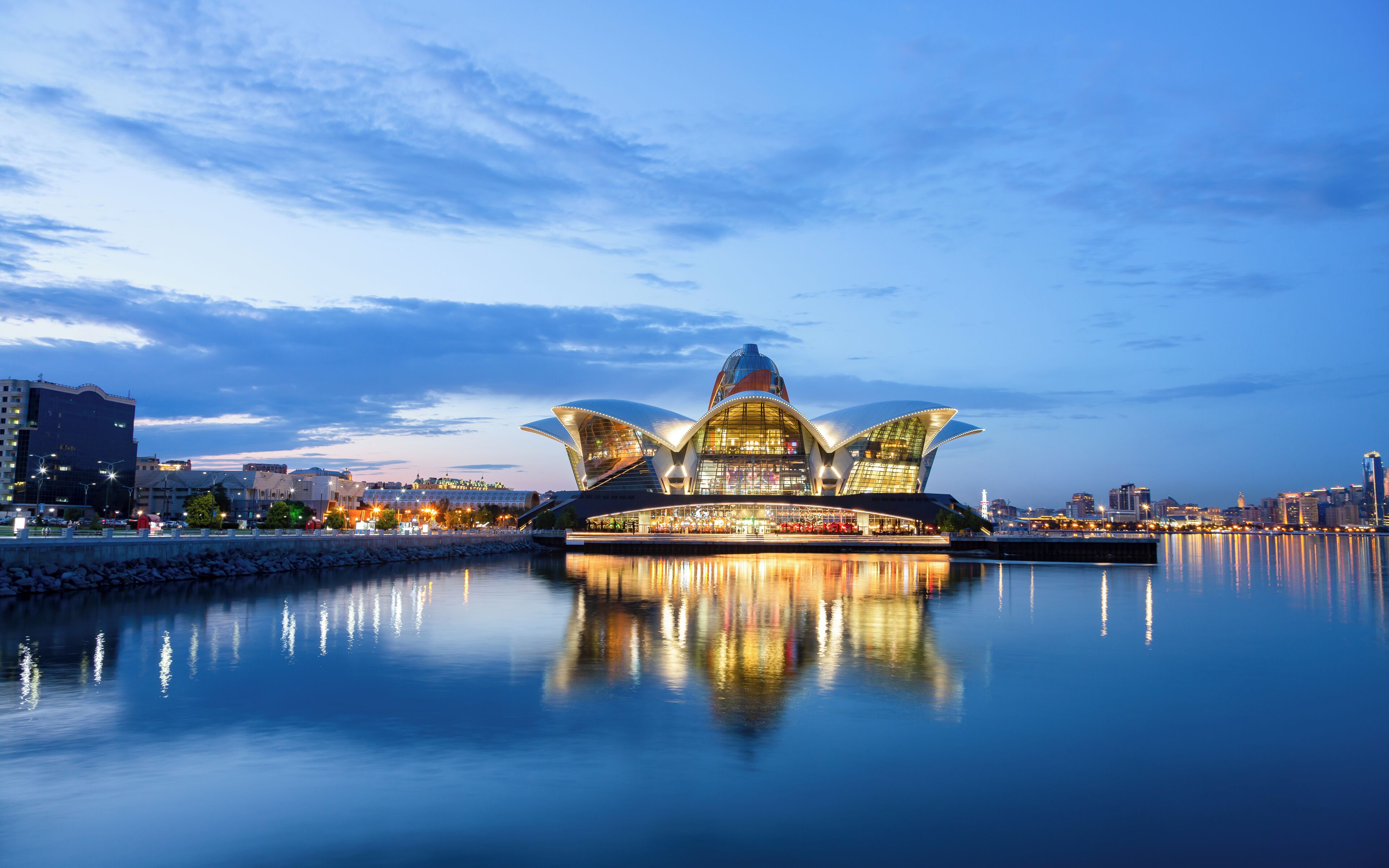 A modern architectural building with a unique lotus-shaped design, situated by a calm body of water reflecting its structure.