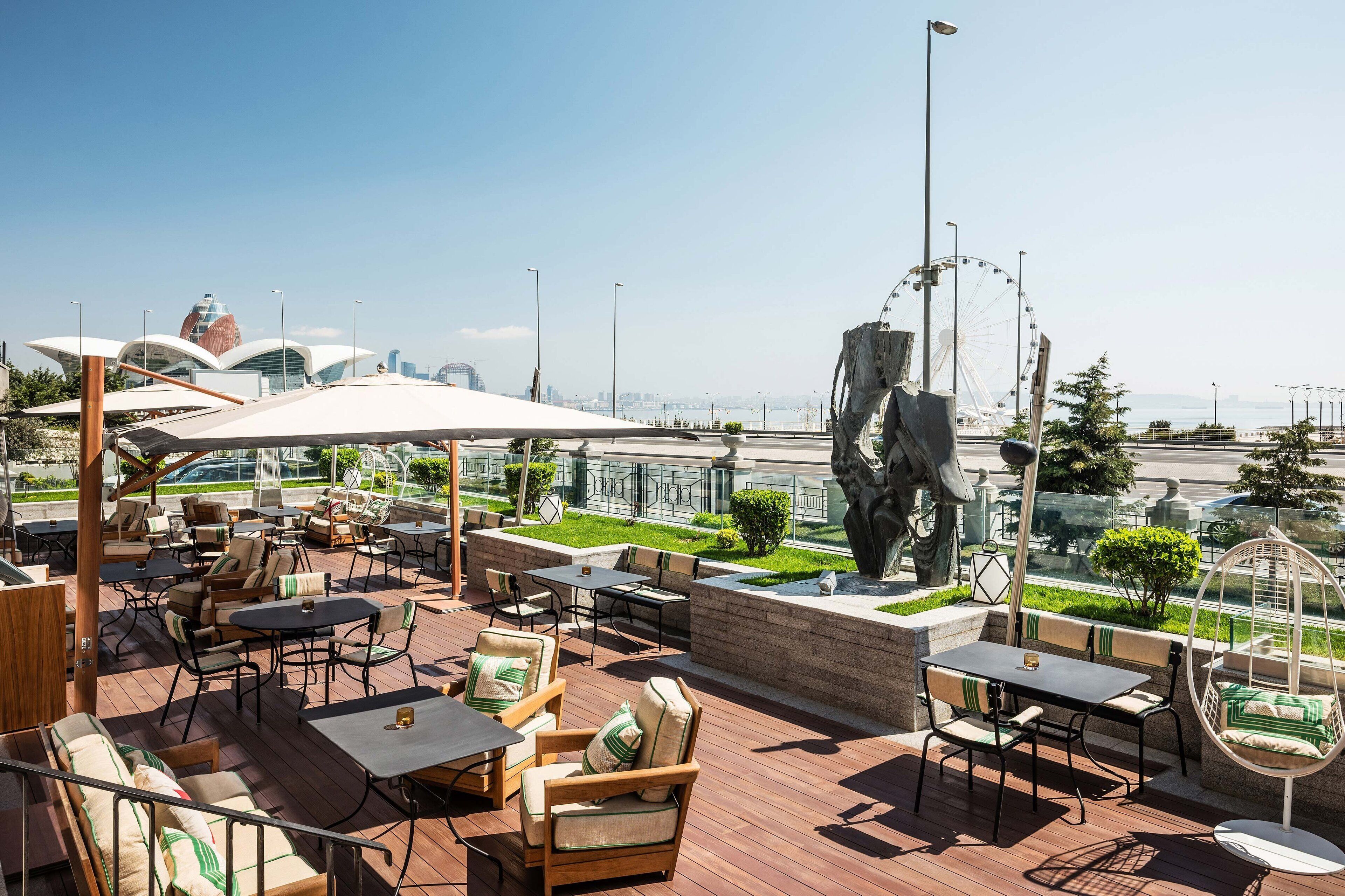 An outdoor seating area with tables and chairs, a large sculpture, and a view of a Ferris wheel and city skyline.