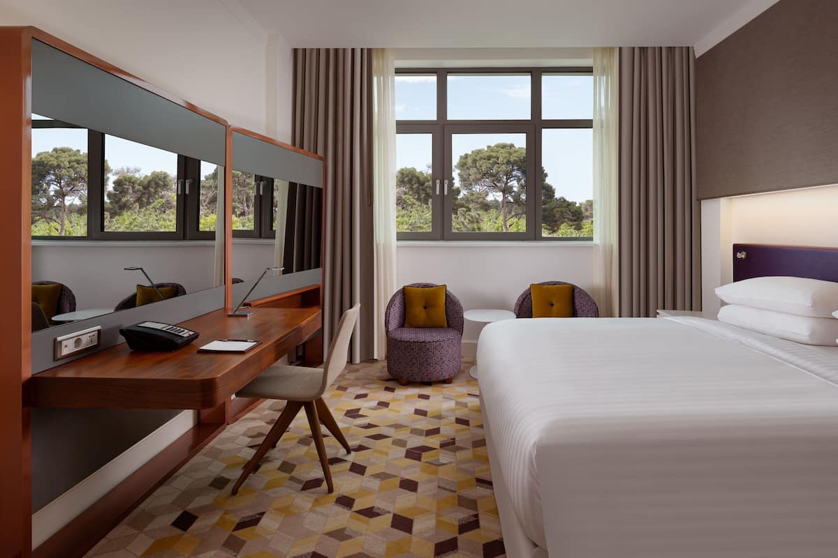 A hotel room with a large bed, a desk with a chair, a small table, and a large mirror. There are two windows with curtains, and a view of trees outside.