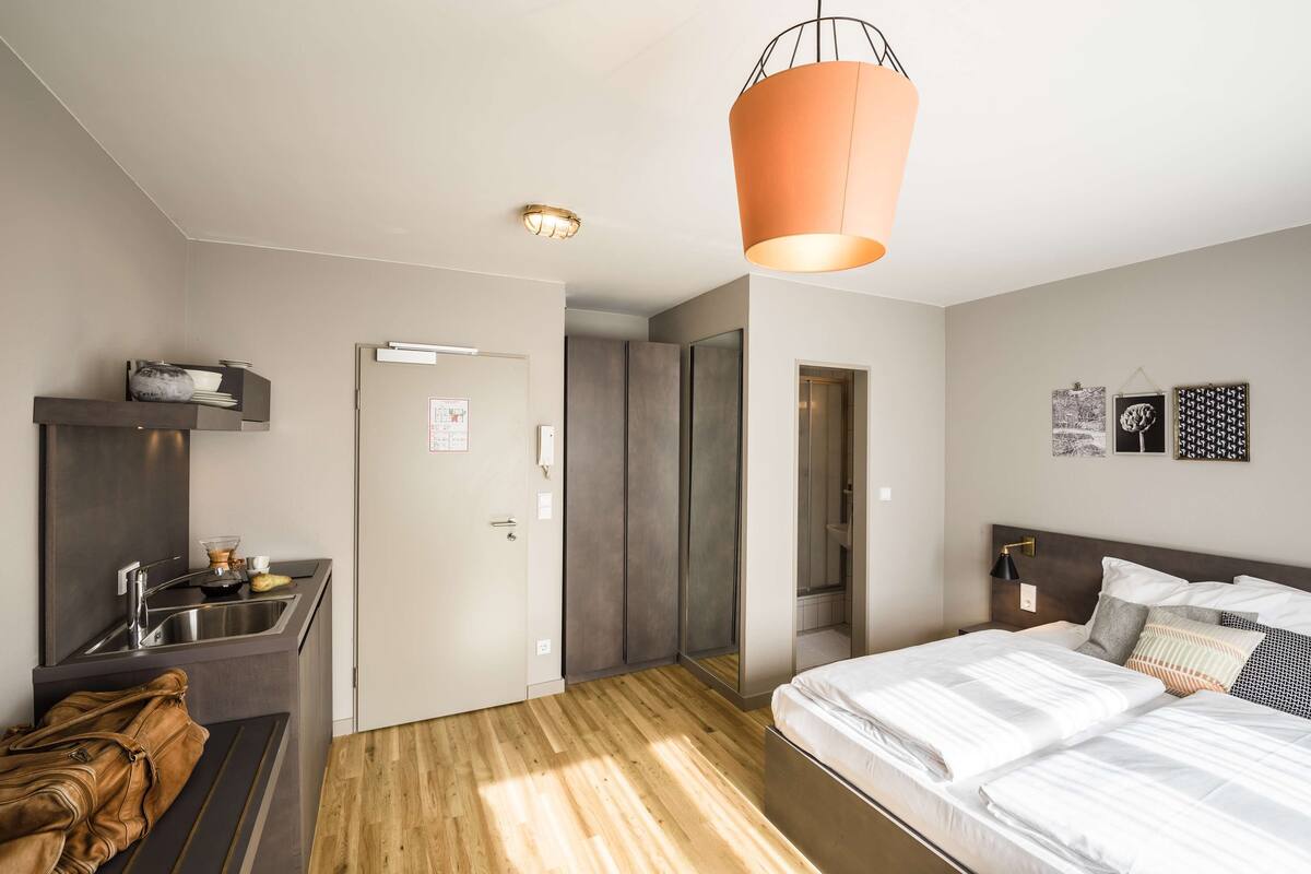 A modern hotel room with a wooden floor, a bed with white linens, a small kitchenette, and a hanging orange pendant light.