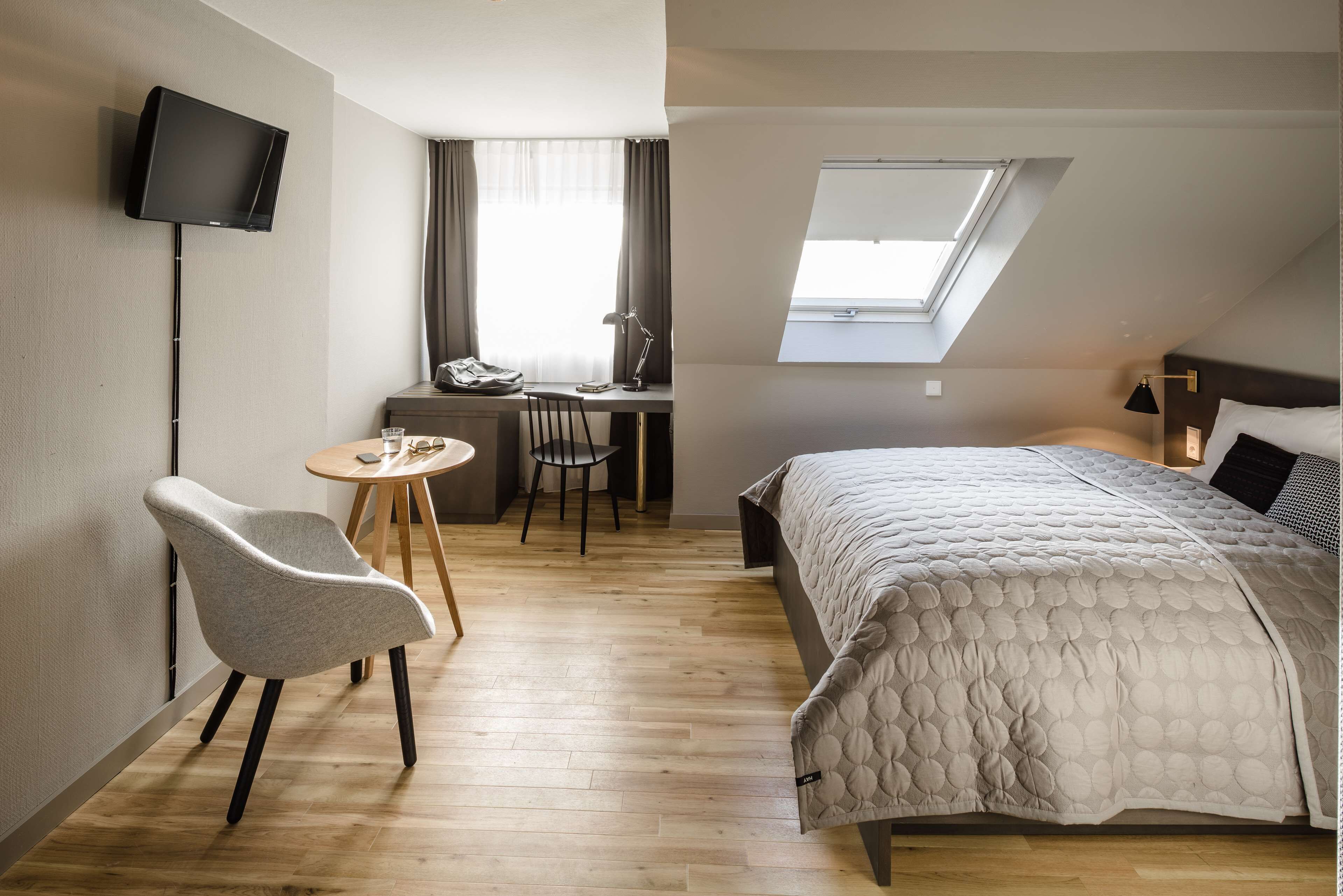 Habitación de hotel moderna con cama, escritorio con silla, televisor montado en la pared y tragaluz.