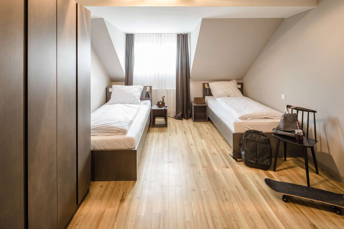 A small, modern hotel room with two single beds, a wooden floor, a desk with a chair, a small table with a cup, and a skateboard.