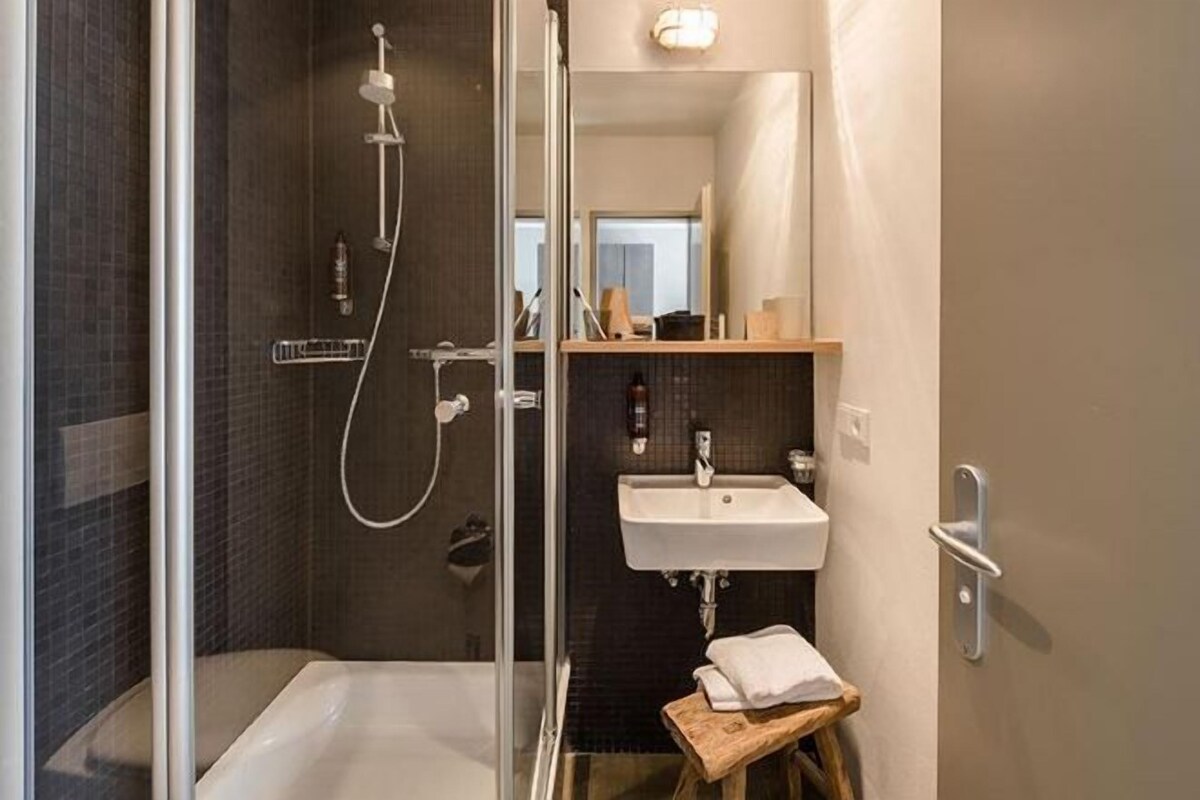 Un baño moderno con ducha envuelta en vidrio, un lavamanos y un taburete de madera.