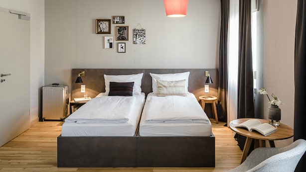 Una cama doble con cabecera oscura, dos mesitas de noche con lámparas, una pequeña mesa redonda con un libro y una silla.
