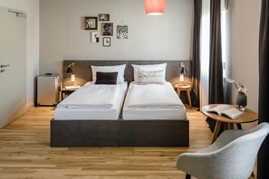 A double bed with a dark headboard, two bedside tables with lamps, a small round table with a book, and a chair. - Bold Hotel Frankfurt Messe (Frankfurt)