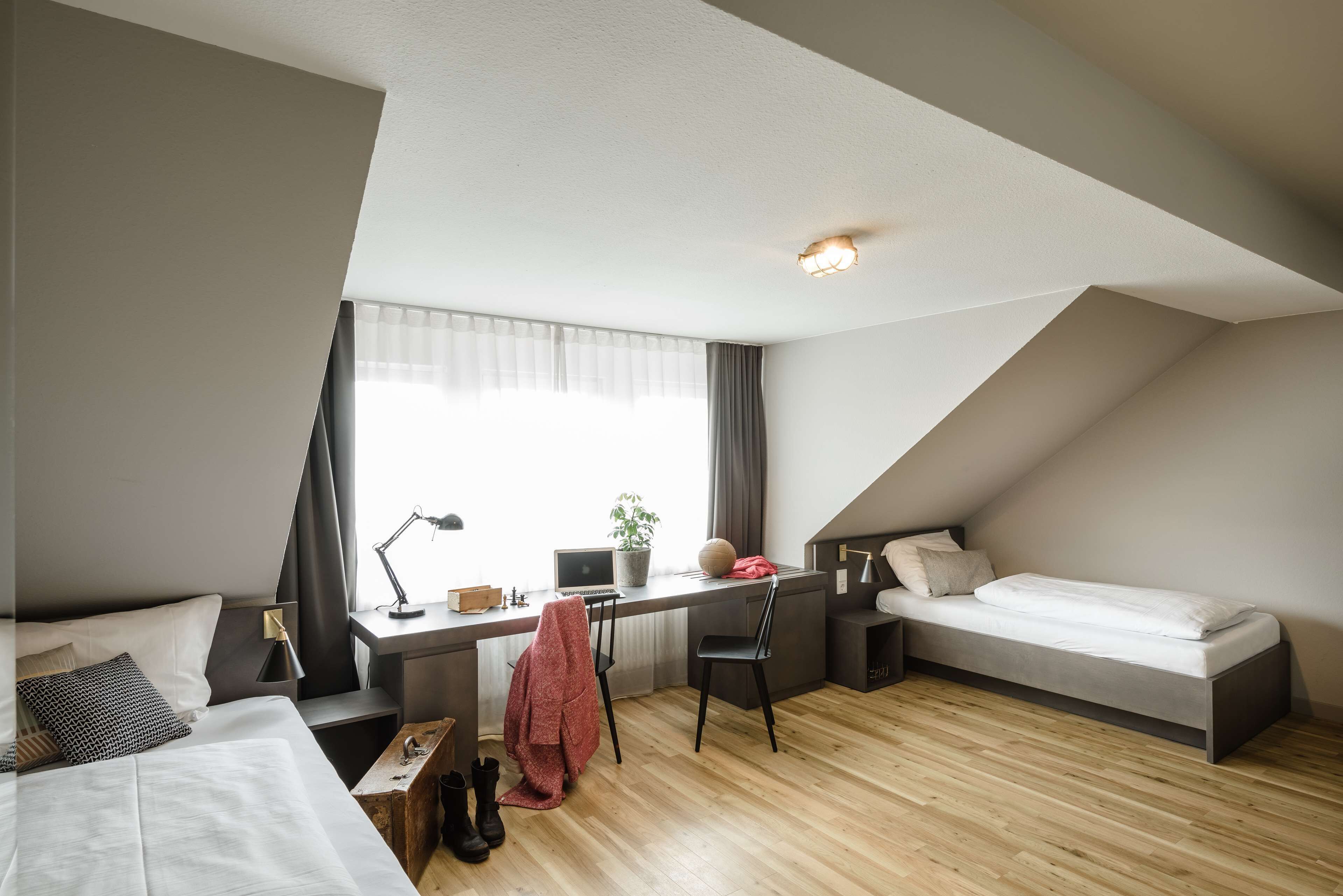A modern, compact room with a bed, desk, and chair, featuring a sloped ceiling and a large window.