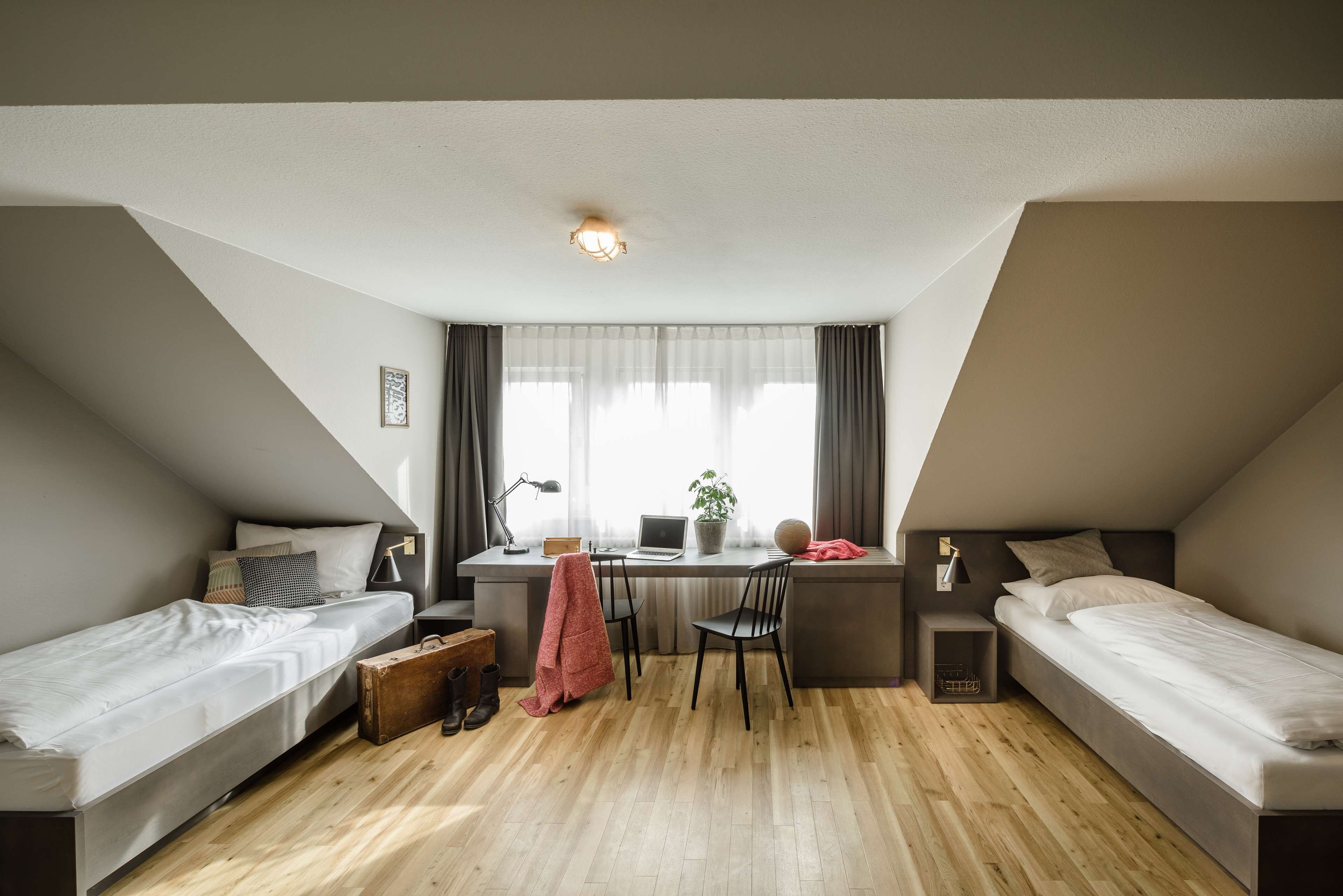 Quarto compacto e triangular com duas camas de solteiro, uma escrivaninha com um laptop, uma cadeira e uma mesinha.