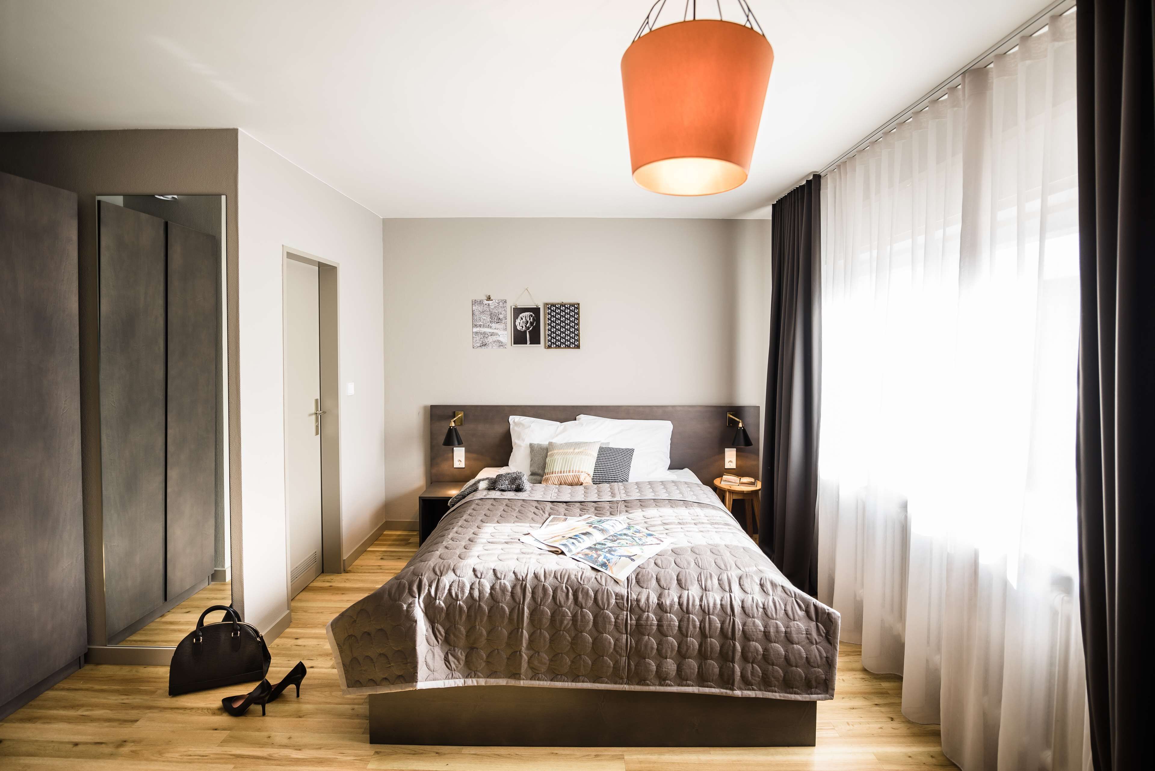 A modern bedroom with a large bed, a hanging light fixture, a wardrobe, and a nightstand with a lamp.