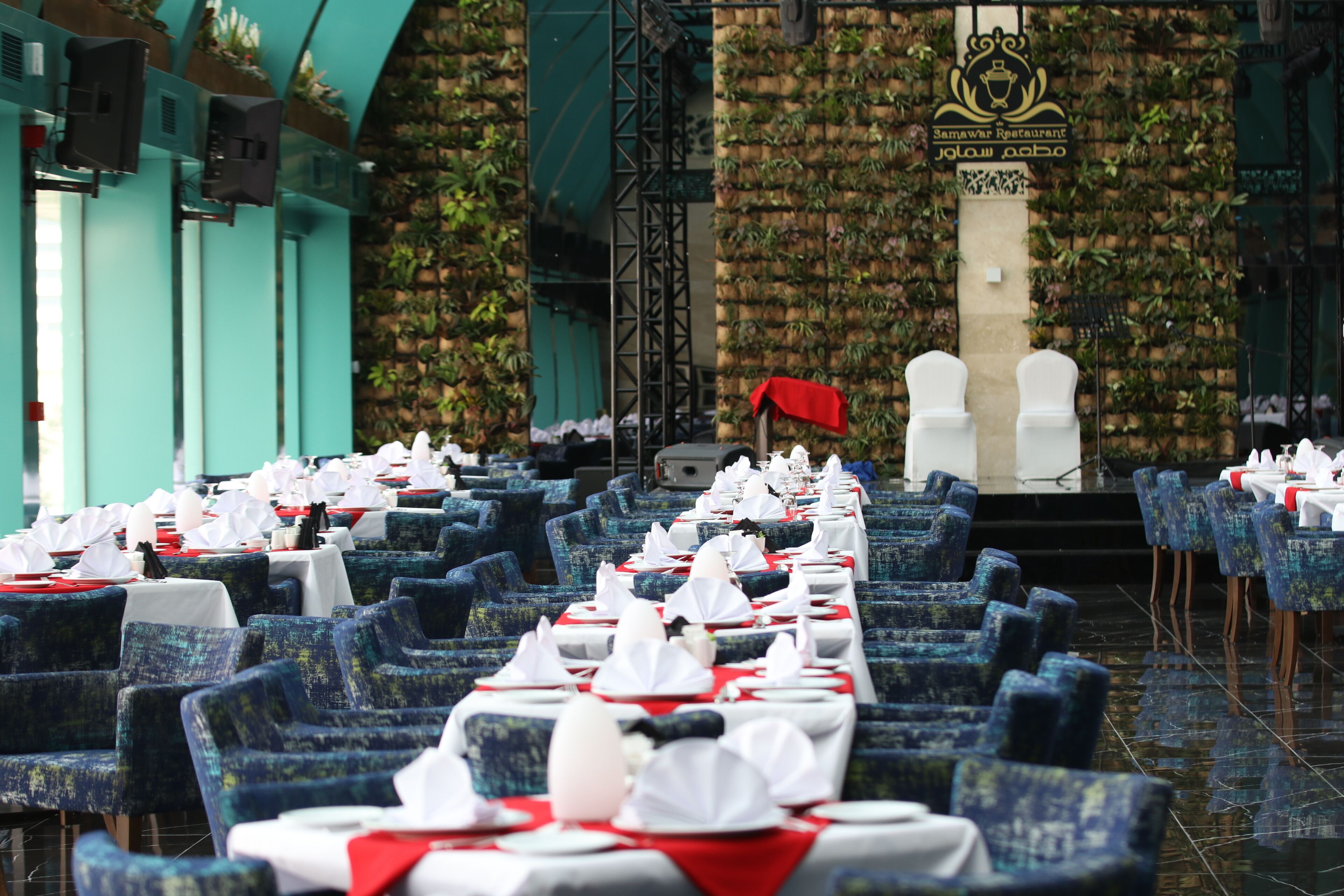 Een restaurant met rijen blauwe beklede stoelen en tafels, gedekt met witte tafelkleden en rode servetten.