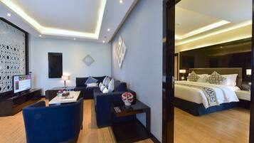 A modern hotel room with a large bed, a seating area with a sofa and chairs, a desk with a computer, and a decorative wall panel.