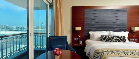 A hotel room with a large bed, a bedside table with a lamp, a chair, a round table with flowers, and a view of the city and water through large windows.