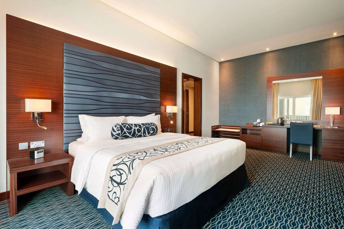 Une chambre d’hôtel avec un grand lit, un bureau et un miroir. Il y a deux tables de chevet avec des lampes.