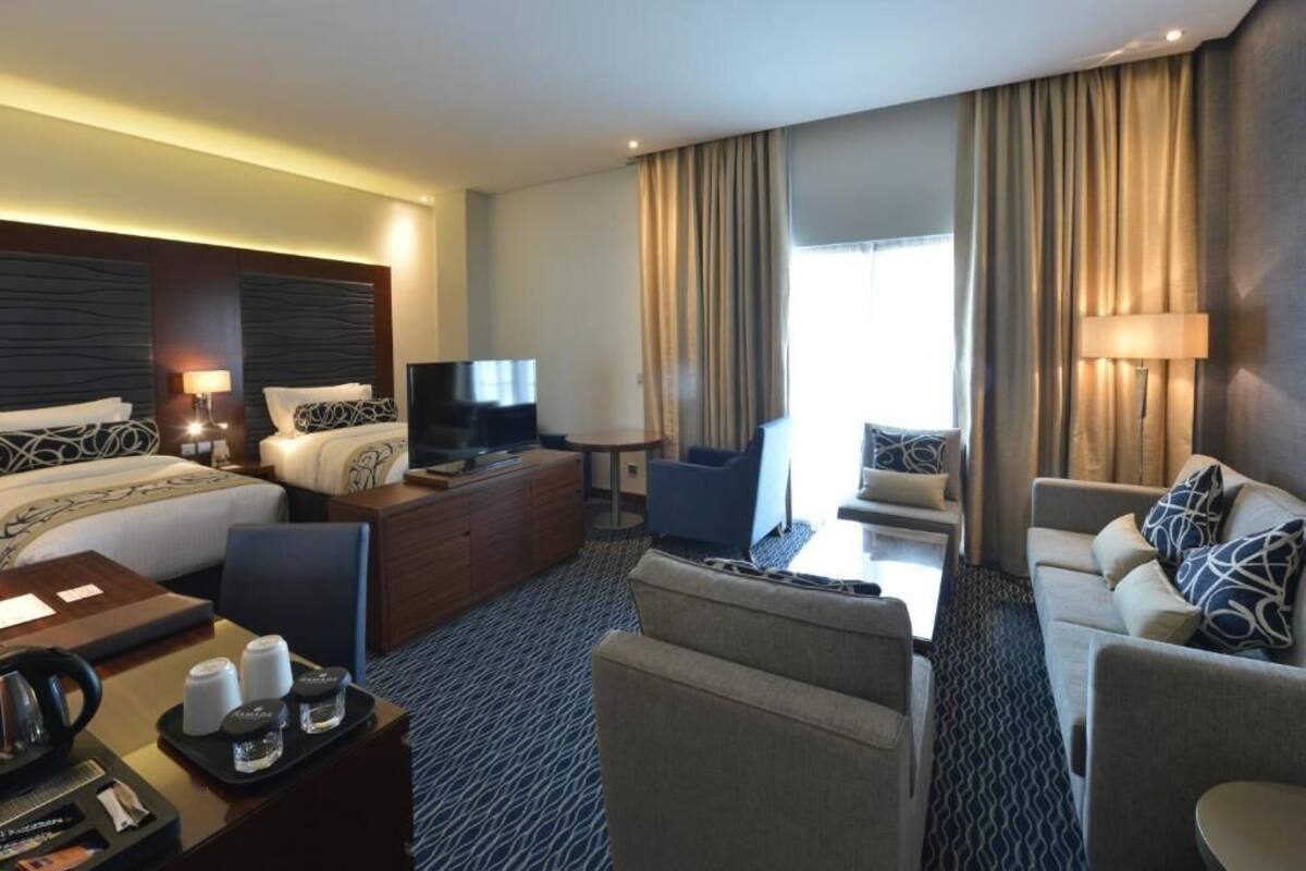 Une chambre d’hôtel comprenant un lit, un canapé, un bureau avec un ordinateur, une télévision et une fenêtre avec des rideaux.