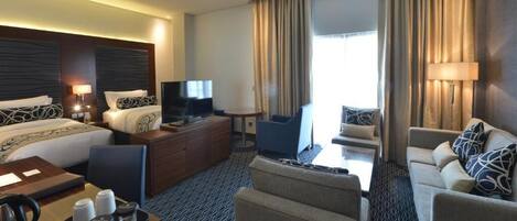 Habitación de hotel con cama, sofá, escritorio con computadora, televisor y ventana con cortinas.